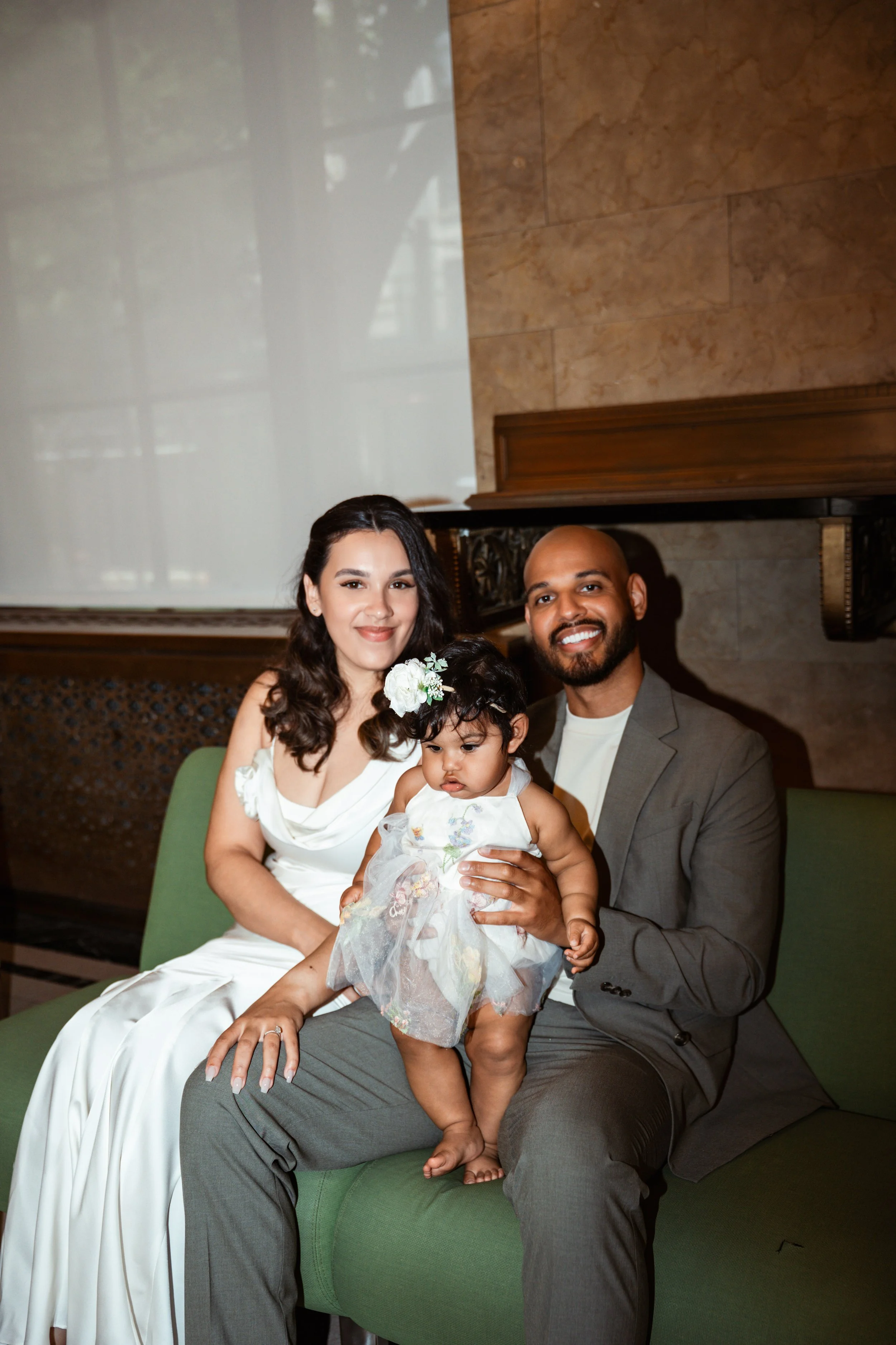 A family portrait of a woman, a man, and a young girl sitting on a green sofa, dressed in formal attire, in an indoor setting with a piano and a marble wall.