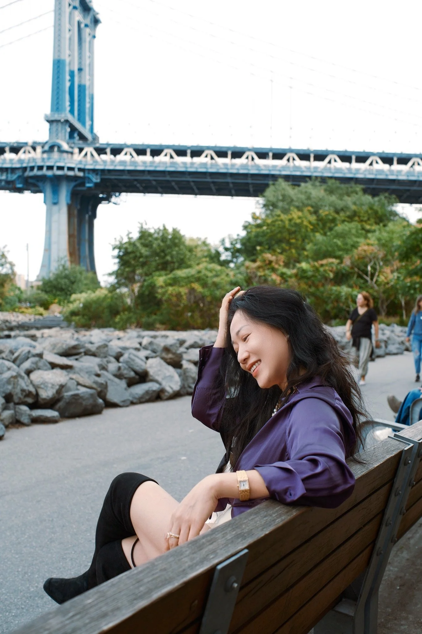 A woman with dark hair and a purple jacket sitting on a wooden bench, smiling and touching her hair in a park near a large bridge, with trees and other people in the background.