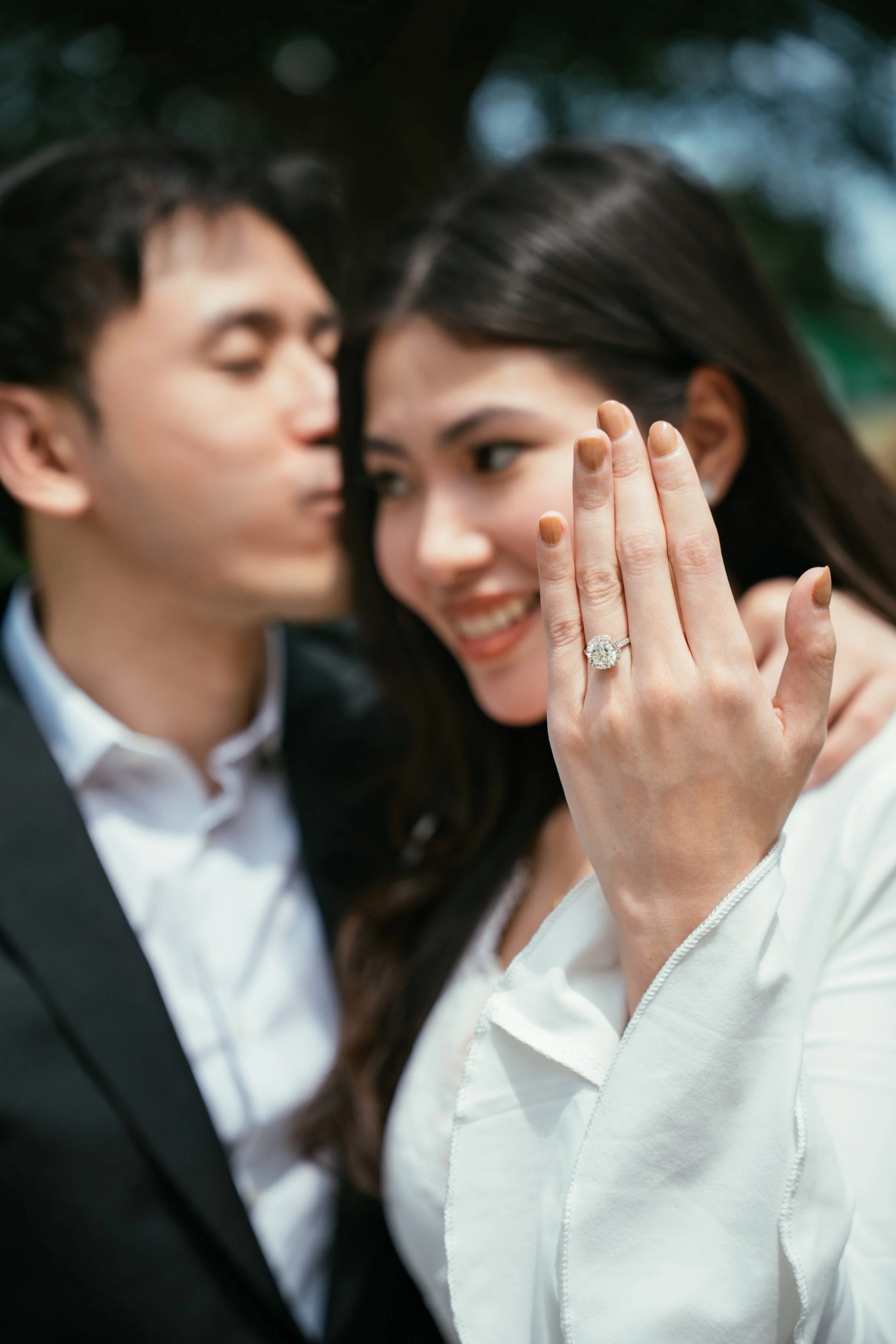 A woman showing her engagement ring to a man, both smiling, outdoors with blurred trees in the background.