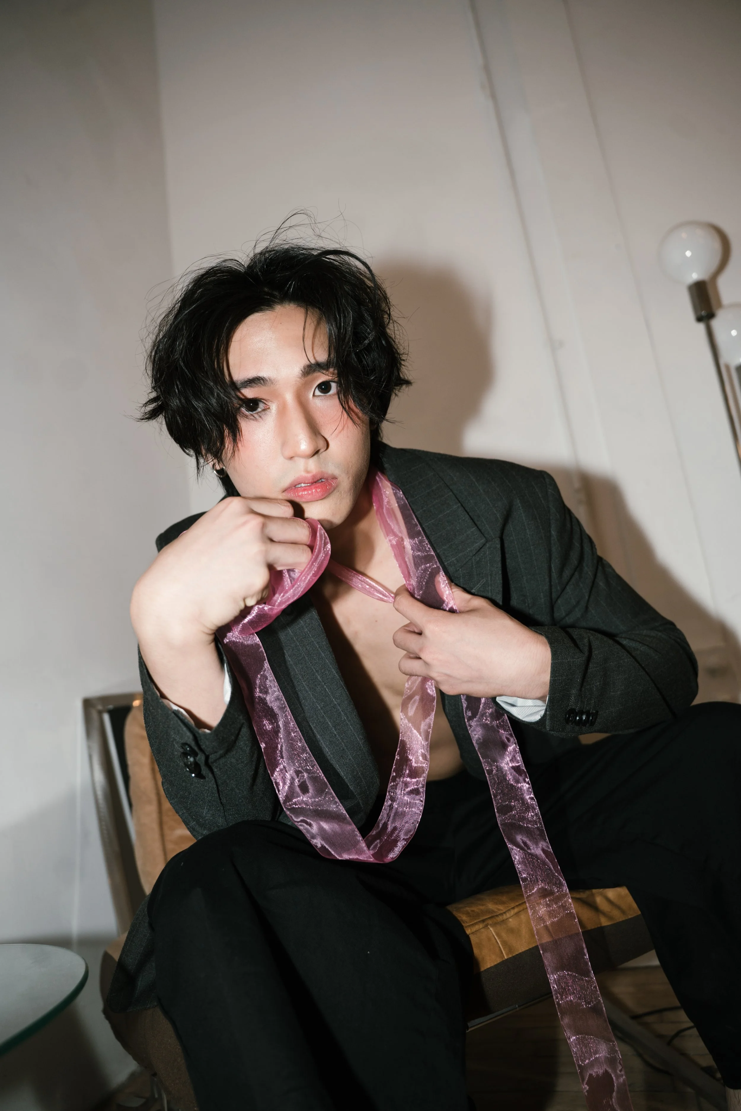 A young man with tousled black hair and fair skin sits on a chair, wearing a dark plaid blazer and a pink satin ribbon around his neck. He is resting his chin on his hand and looking into the camera with a serious expression. The background is white 