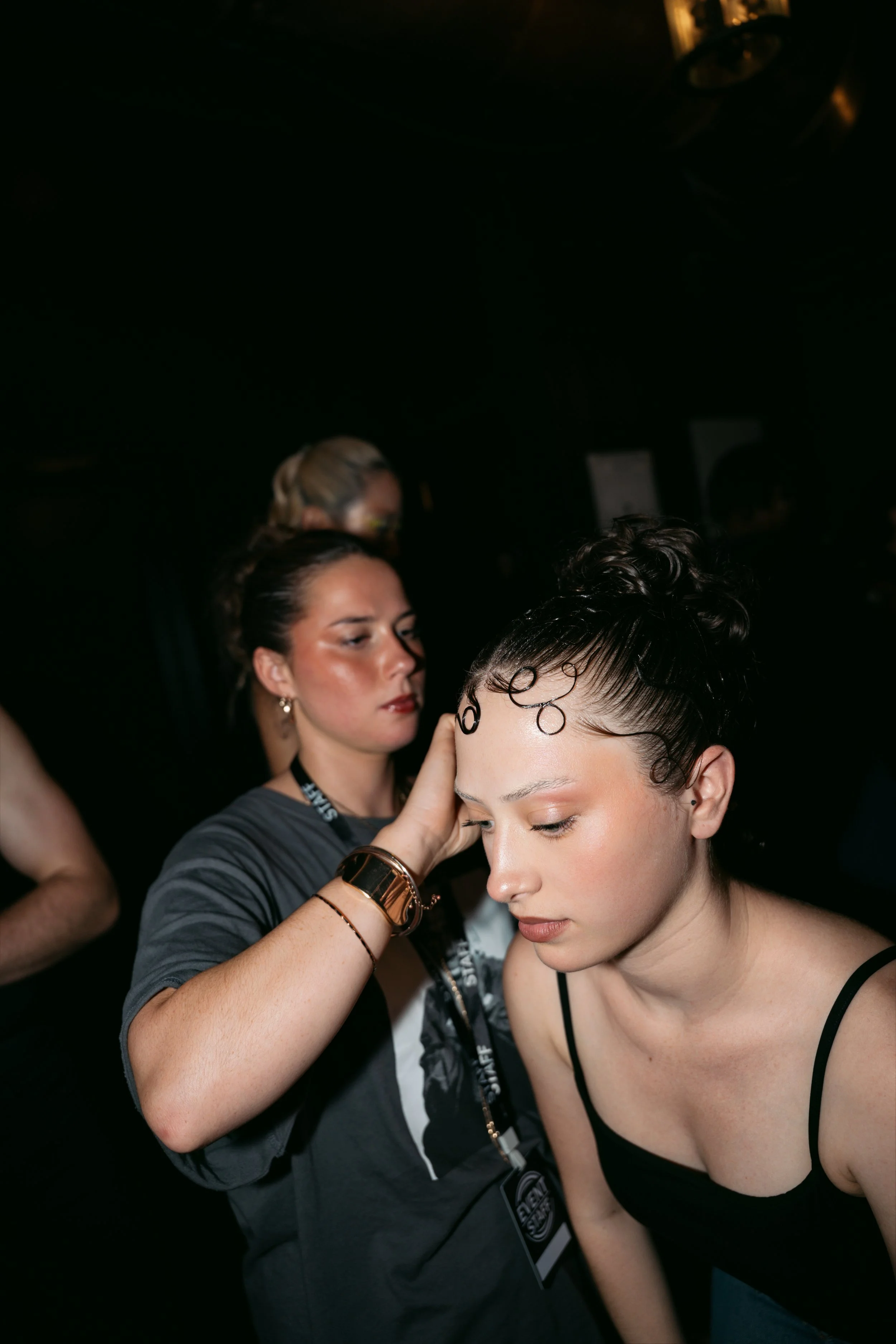 A makeup artist is applying makeup to a young woman with curly hair and a black sleeveless top. The makeup artist is wearing a black lanyard, gold bracelet, and has her hair tied back. The background is dark, and there are other people visible in the