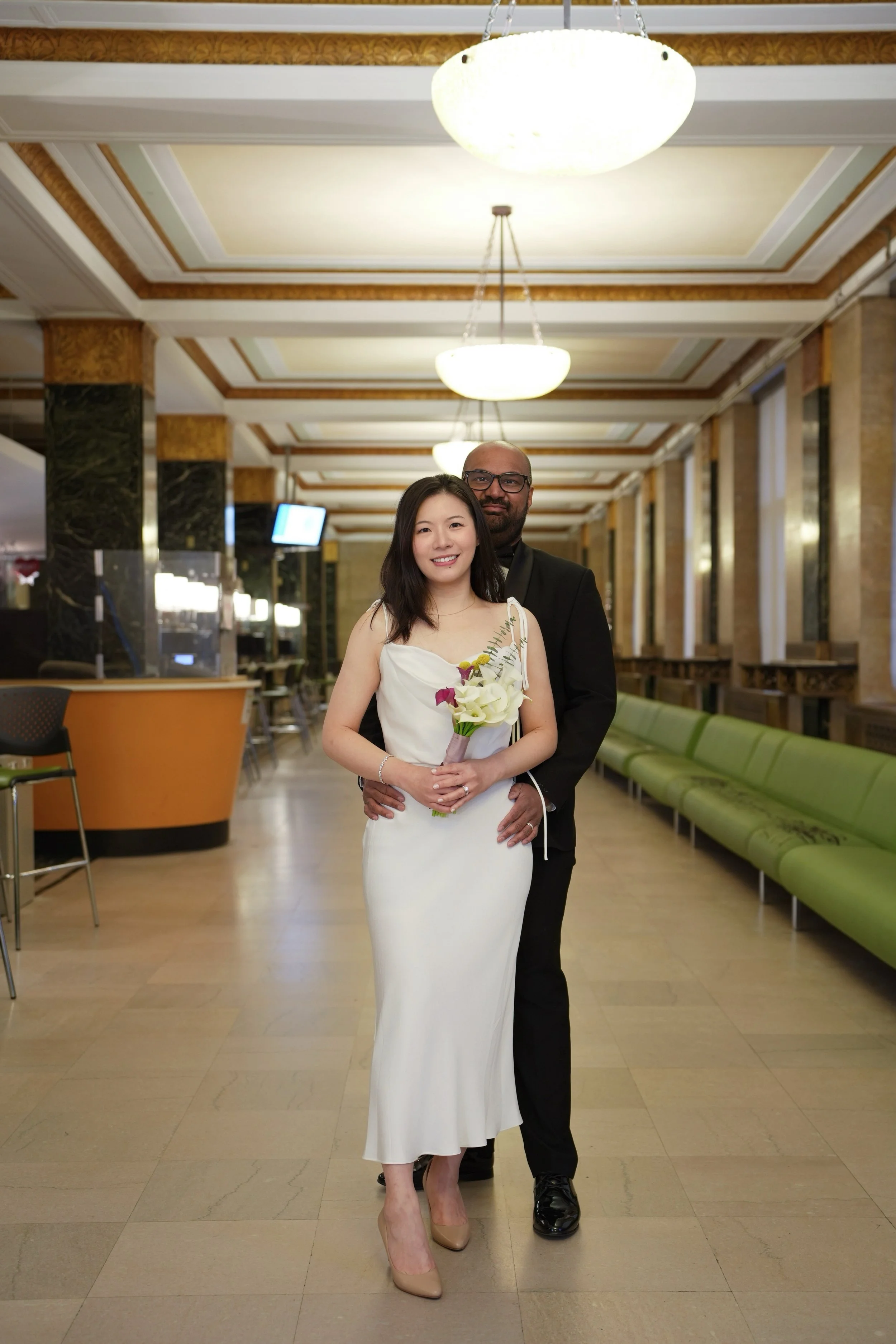 A joyful couple dressed for a wedding, with the woman in a white gown holding a bouquet of white and purple flowers, and the man in a black suit, standing in a warmly lit indoor venue with green seating and decorative lighting fixtures.