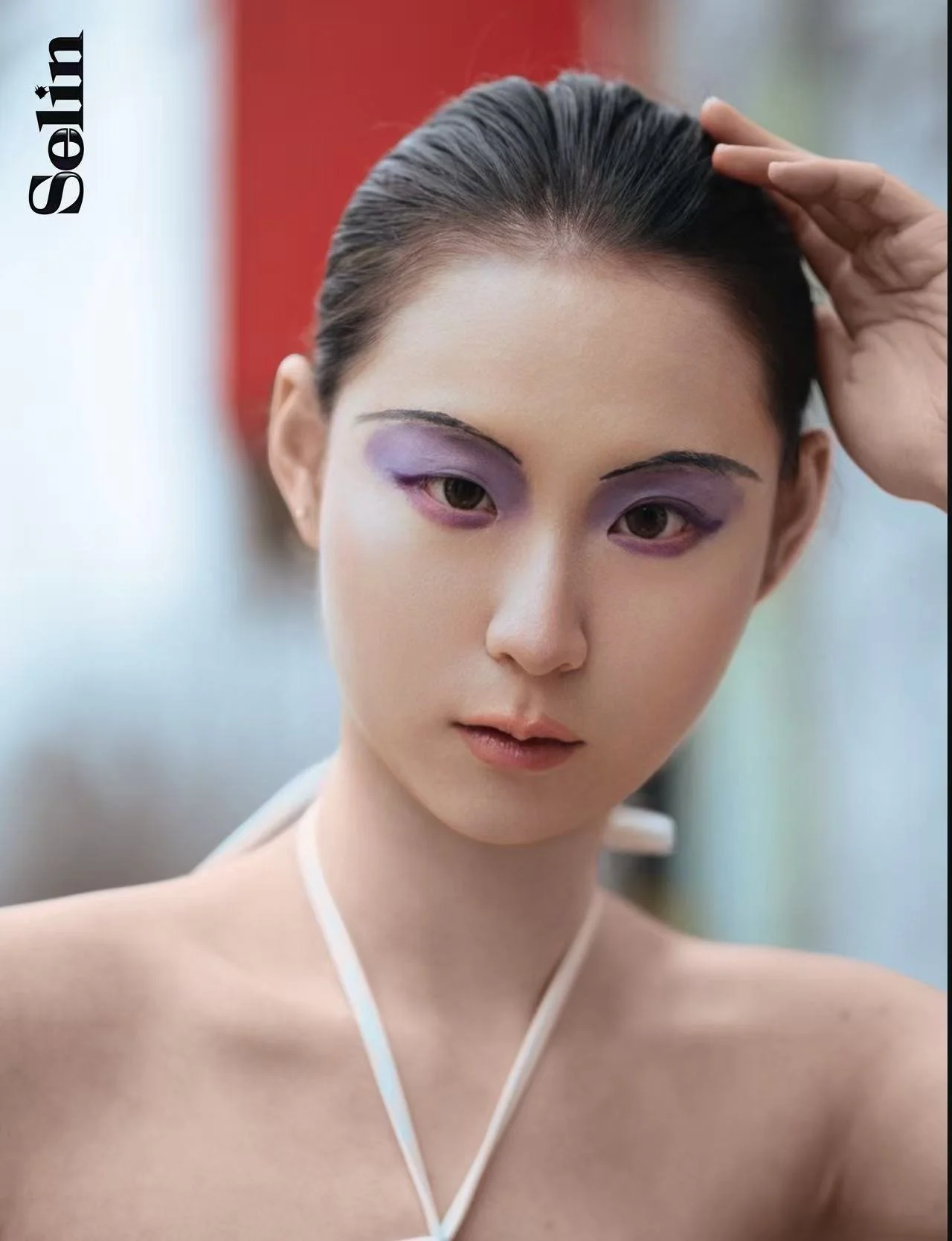 A young woman with dark hair styled back, wearing purple eyeshadow, holding her hand to her head, and looking into the camera, with a blurred background.