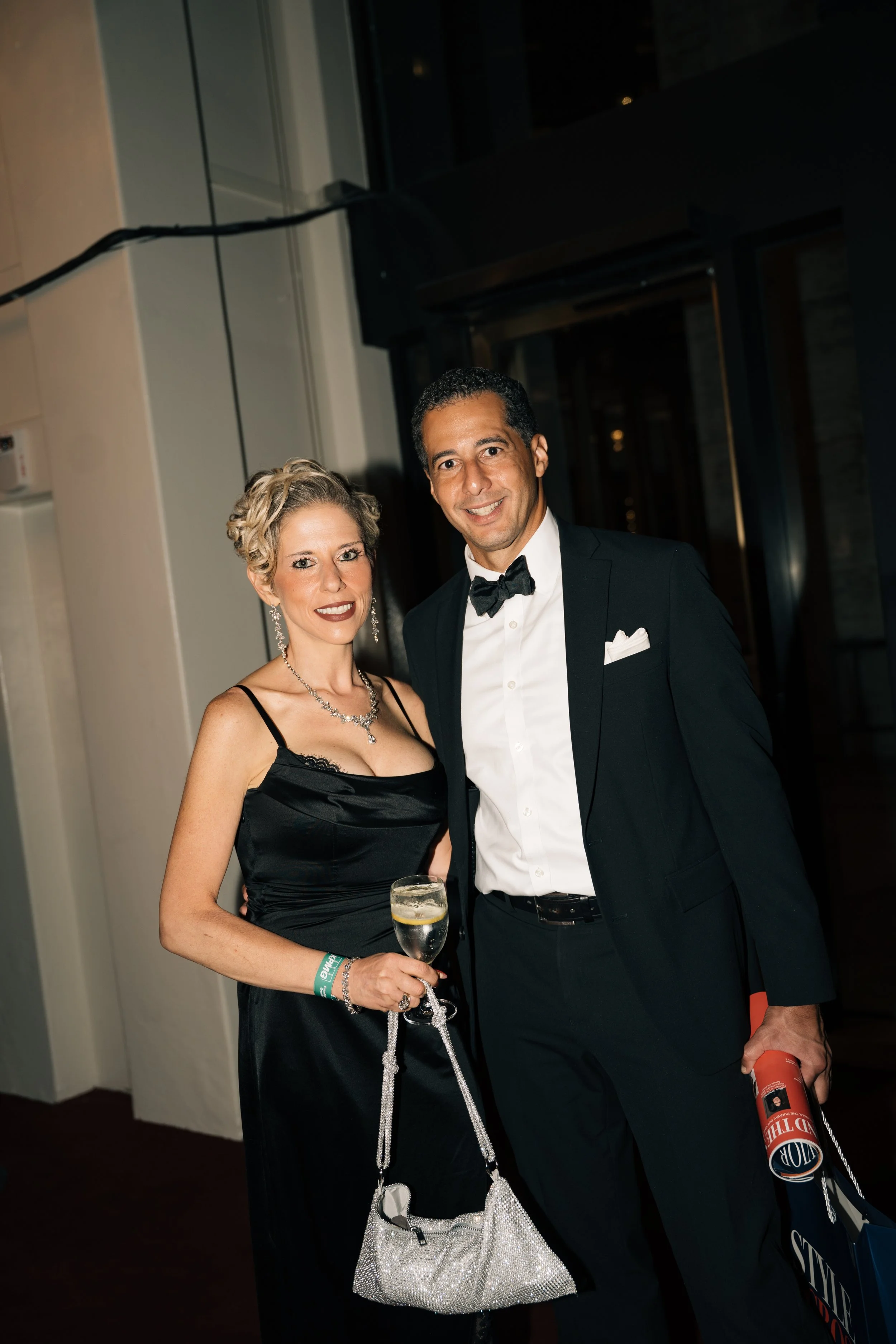A woman in a black dress and a man in a tuxedo posing at a formal event, with the woman holding a glass of wine and wearing jewelry and a silver handbag, and the man holding a rolled-up poster or document.