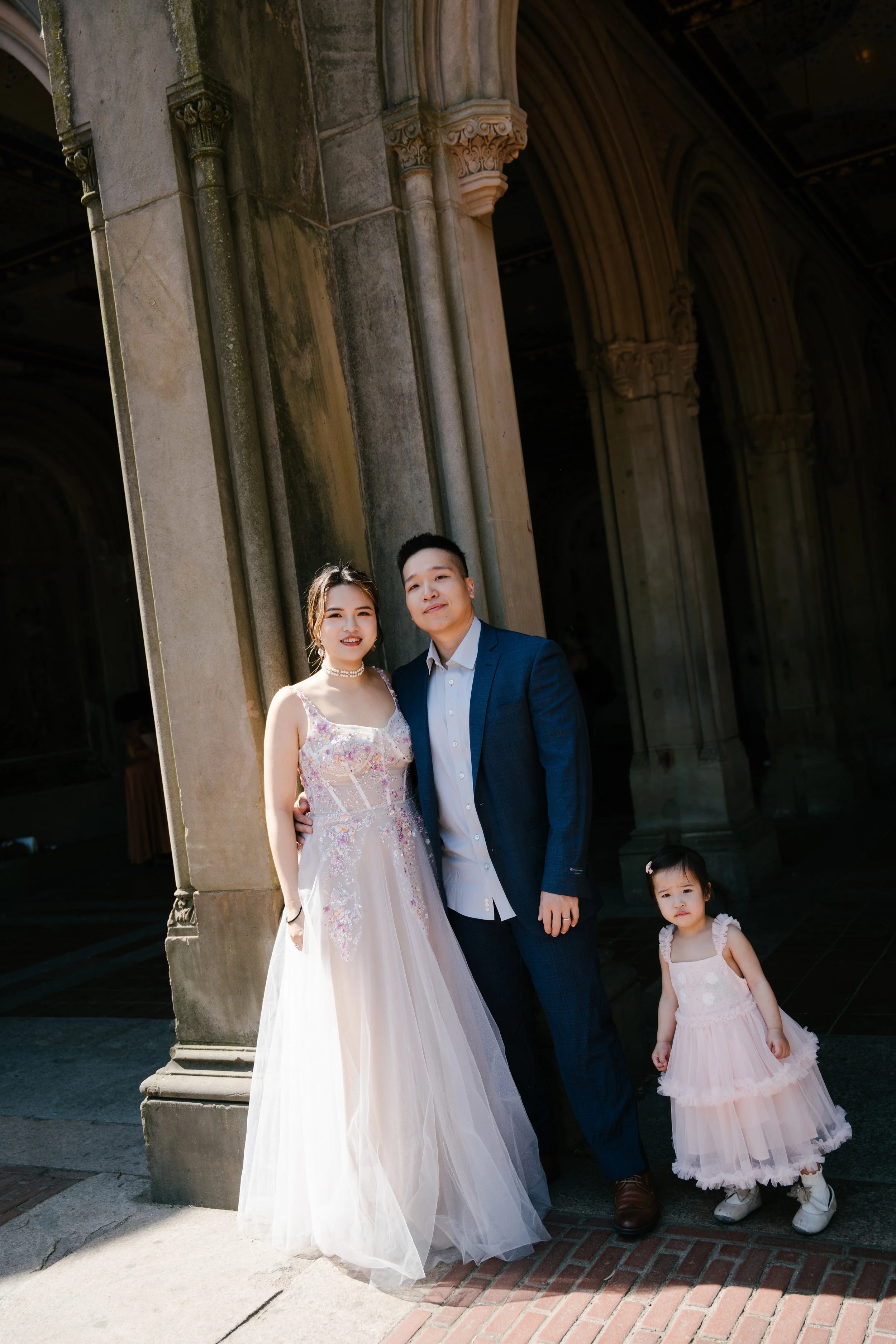 A family of four standing outside a historic stone building with Gothic arches. The woman is wearing a white and pink wedding dress, and the man is dressed in a dark blue suit. There is a young girl in a pink dress with a tulle skirt standing beside 