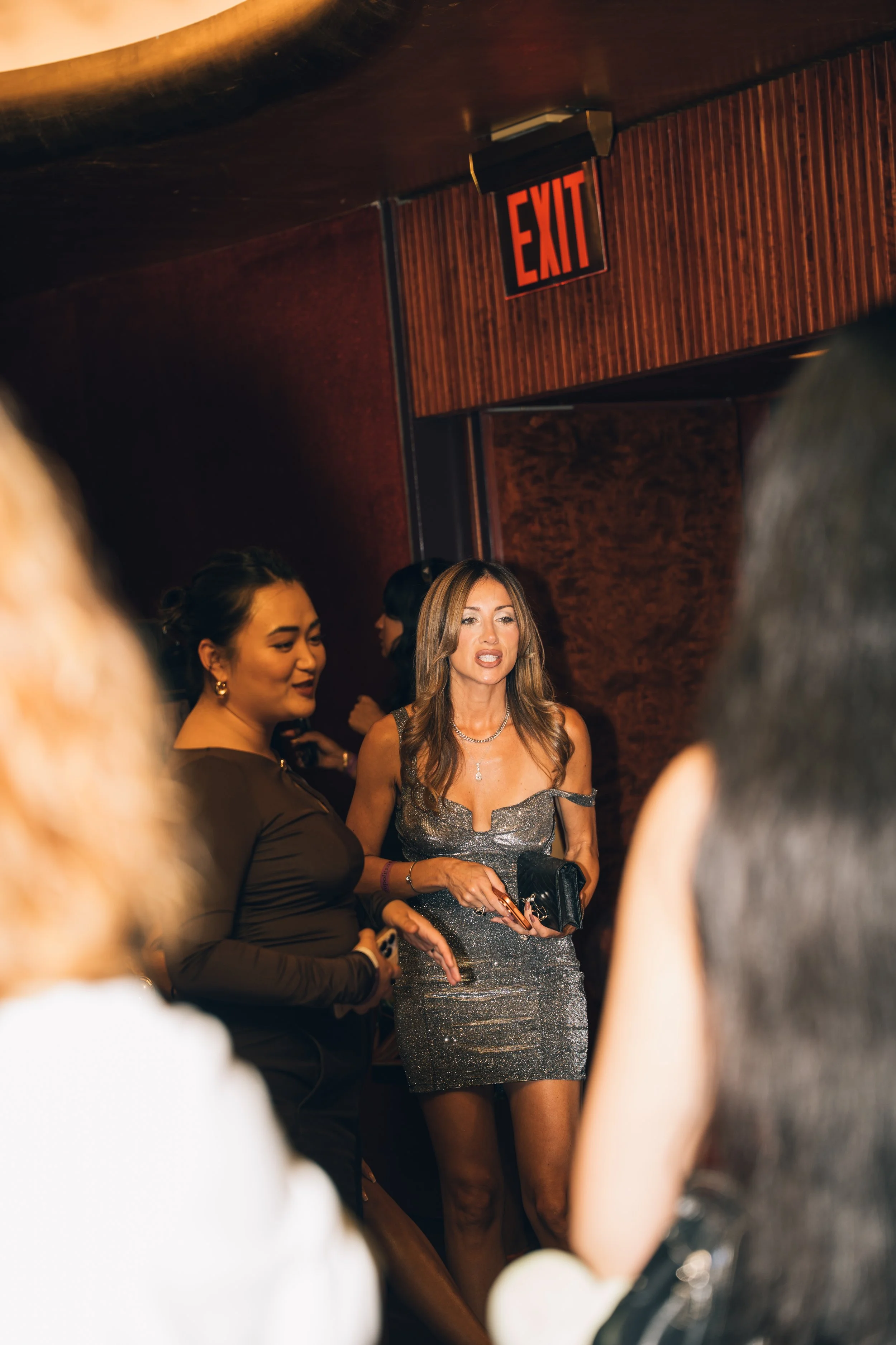 Women dressed in evening wear at an indoor event with a wood-paneled wall and an illuminated exit sign in the background.