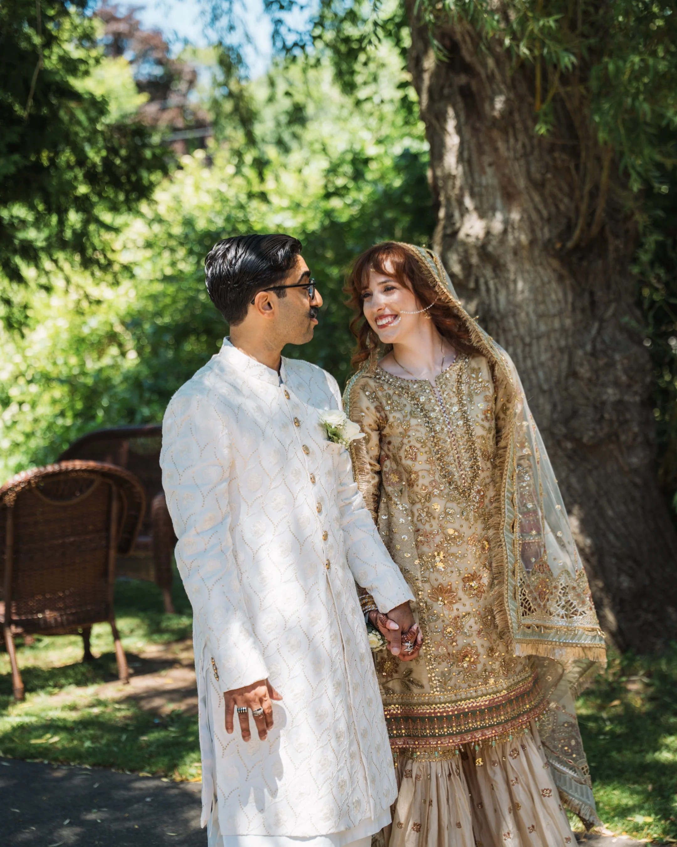A couple dressed in traditional South Asian wedding attire holding hands outdoors near a large tree, with lush green foliage and a wicker chair in the background.