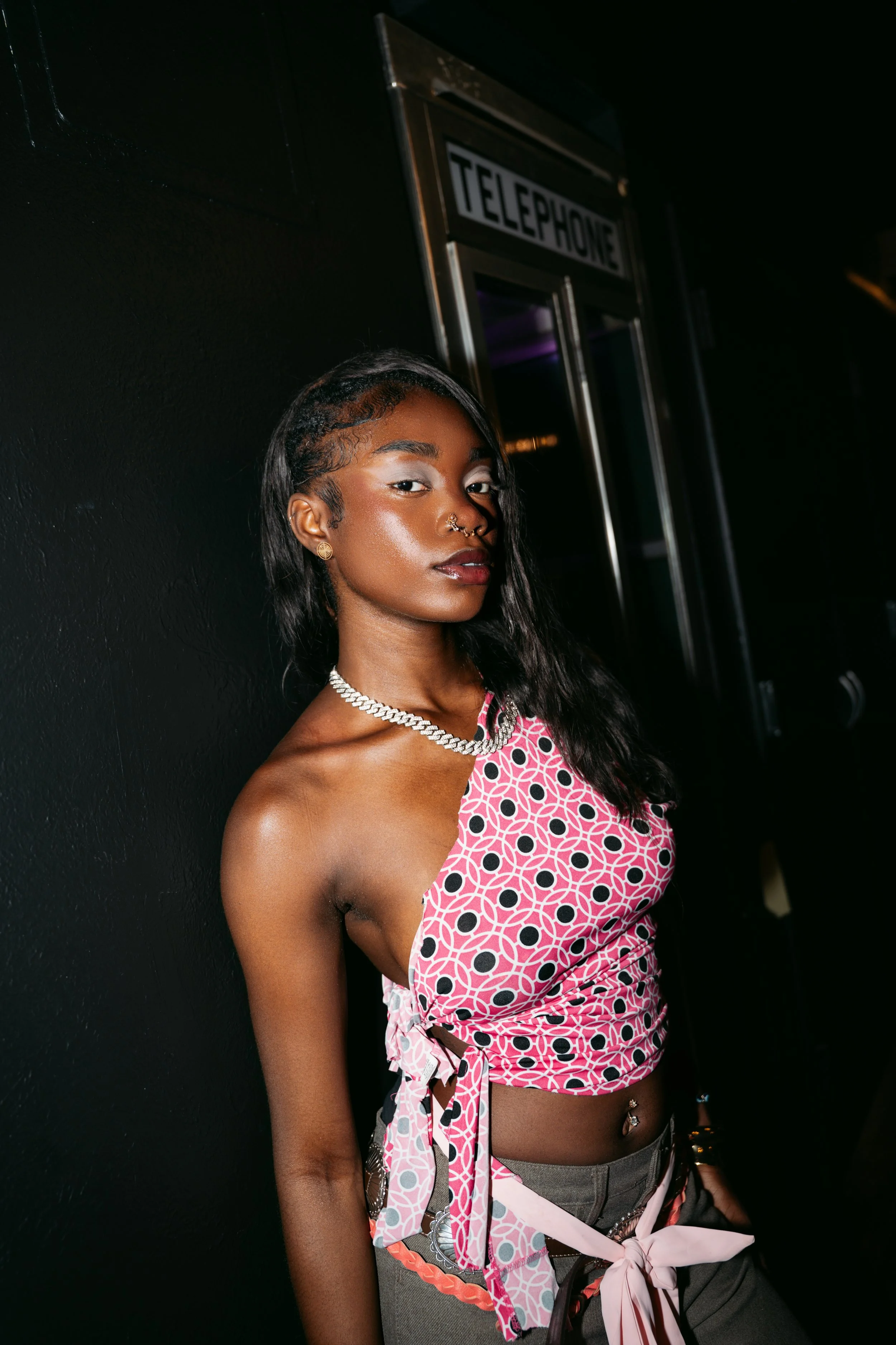 A young woman with dark hair styled in waves, wearing gold earrings, a gold chain necklace, and a septum piercing, posing against a black wall with a vintage telephone booth in the background.