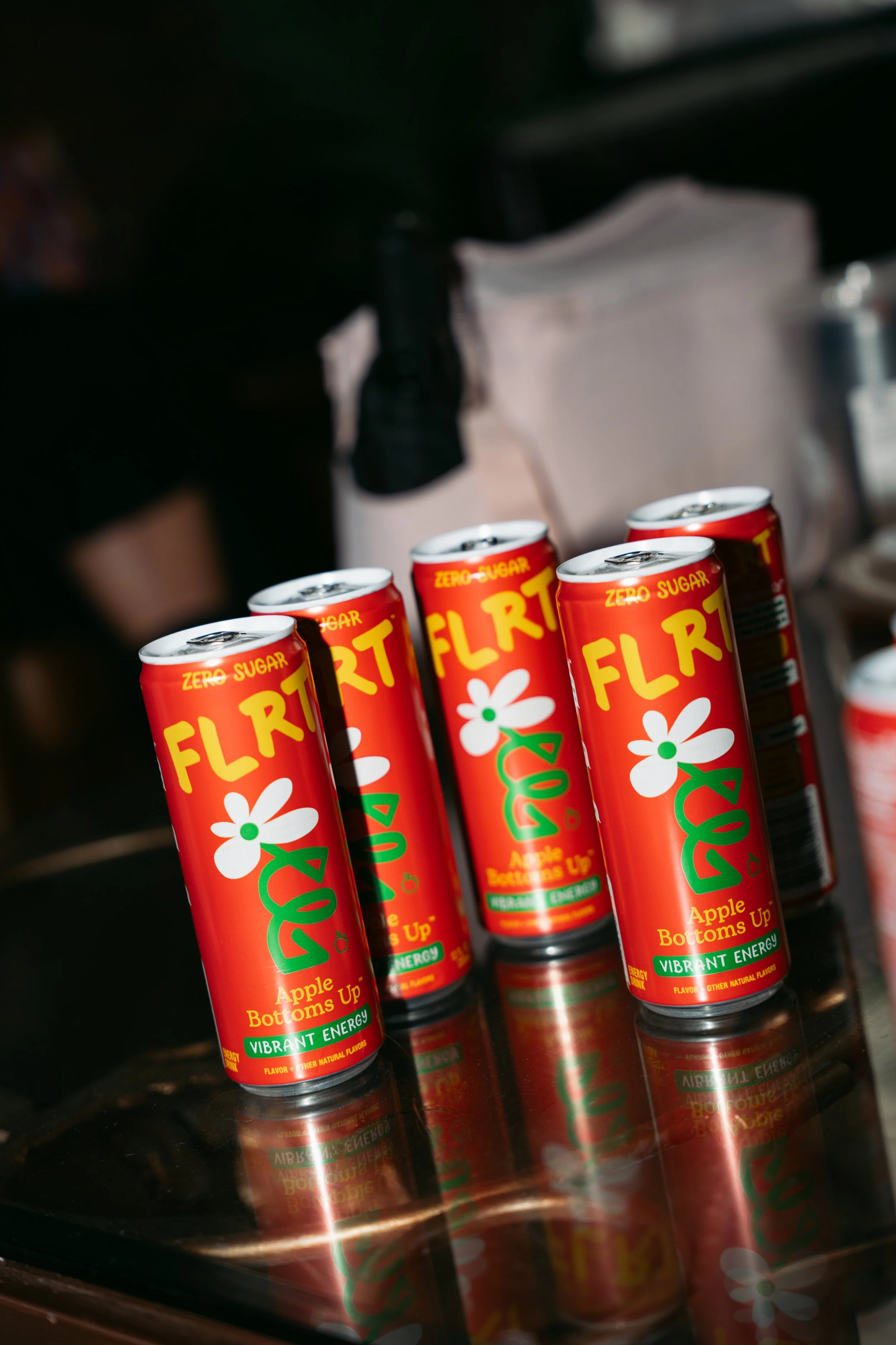Several cans of Flirt zero sugar apple flavored beverage with vibrant orange and green packaging arranged on a reflective surface.