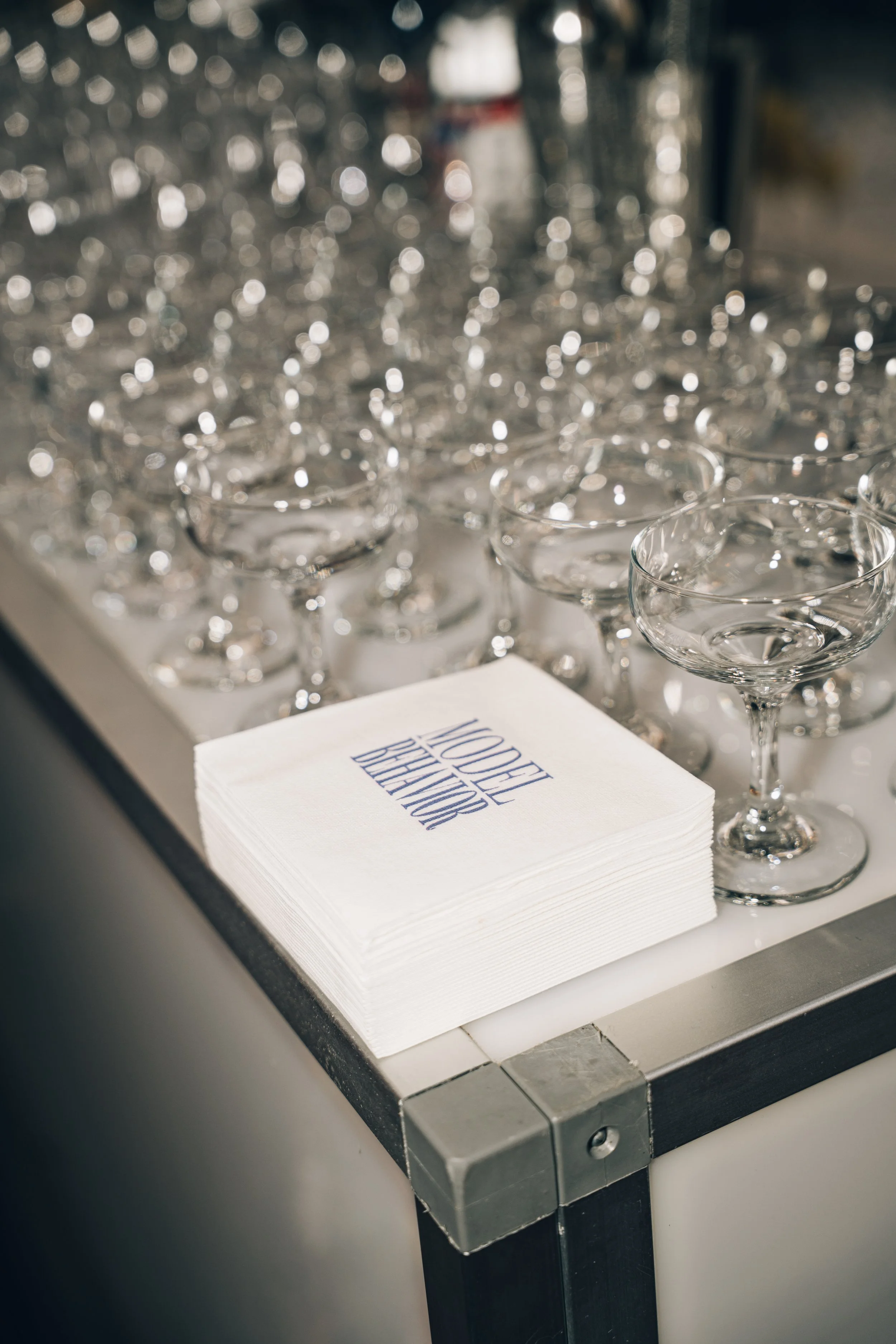 A stack of white paper napkins with the words 'WOODFELD BREWERY' printed on top, placed on a metal table near rows of empty cocktail glasses, with a blurred background showing a glass jar and some other items.