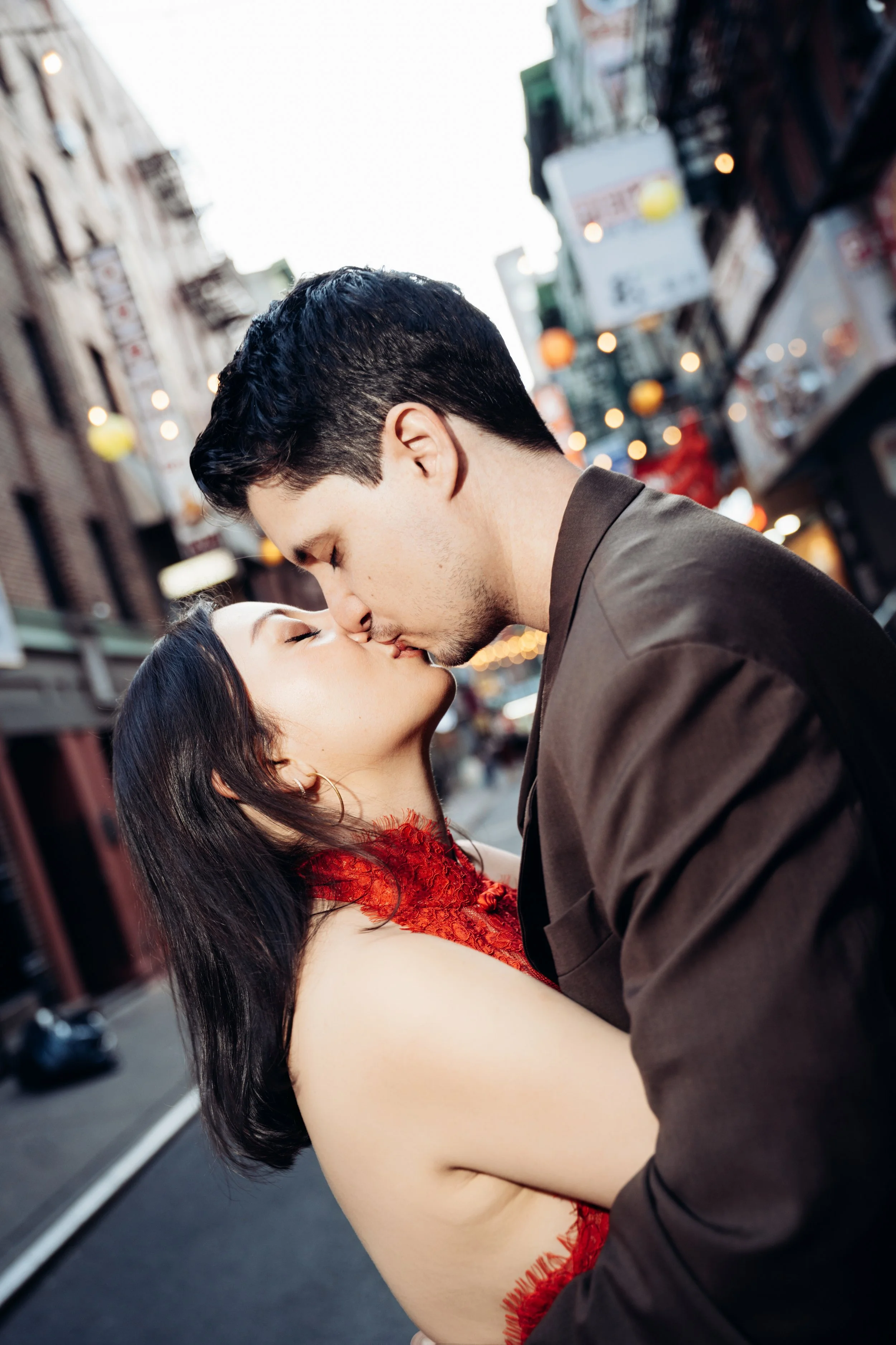 A woman in a red dress and a man in a brown suit kiss on a city street with blurred lights and signs in the background.