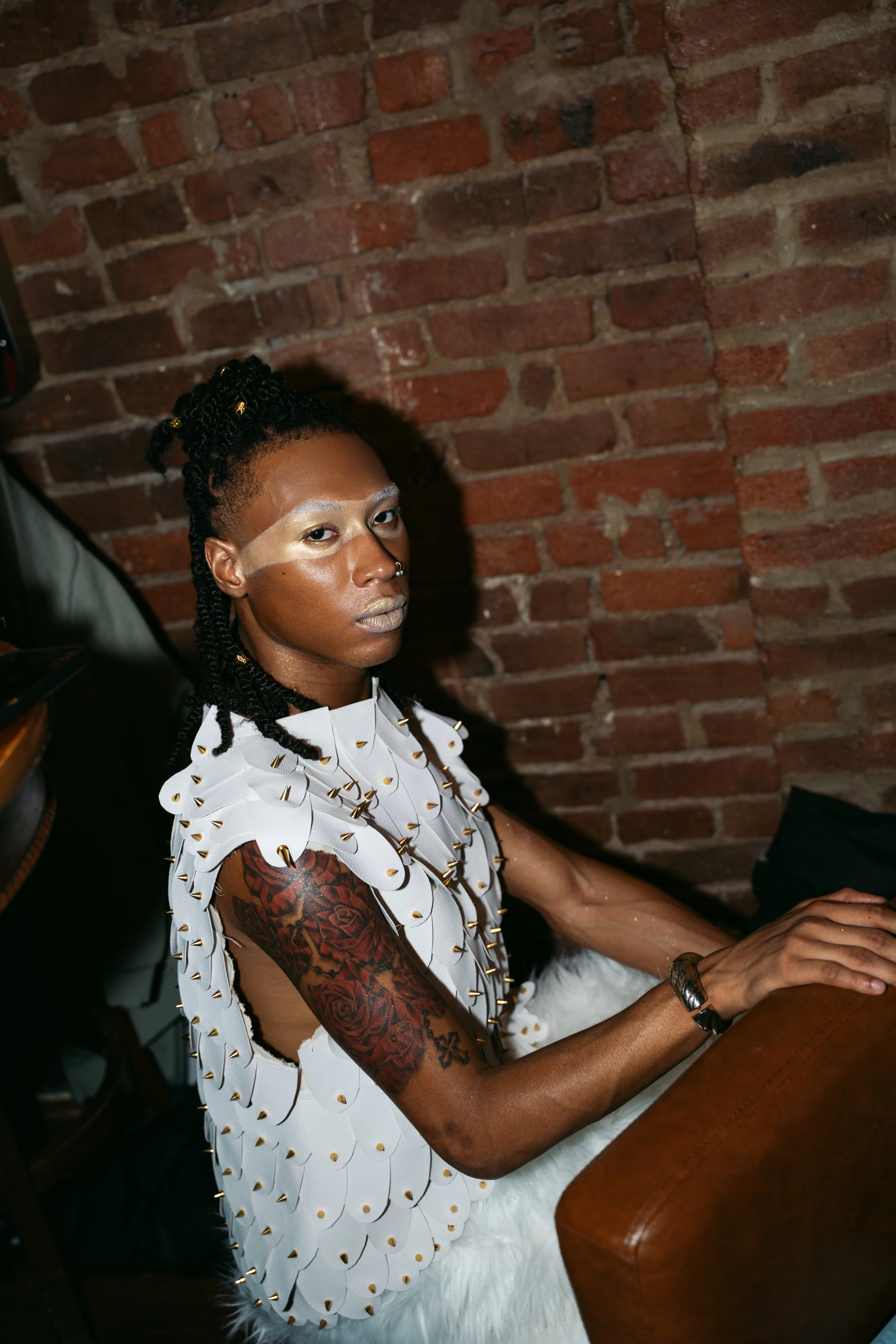 A person with dark skin, styled in twists with gold beads, and arm tattoos, sitting at a table with a brick wall background. They wear a white vest with gold spikes, makeup with metallic accents, and a nose ring.