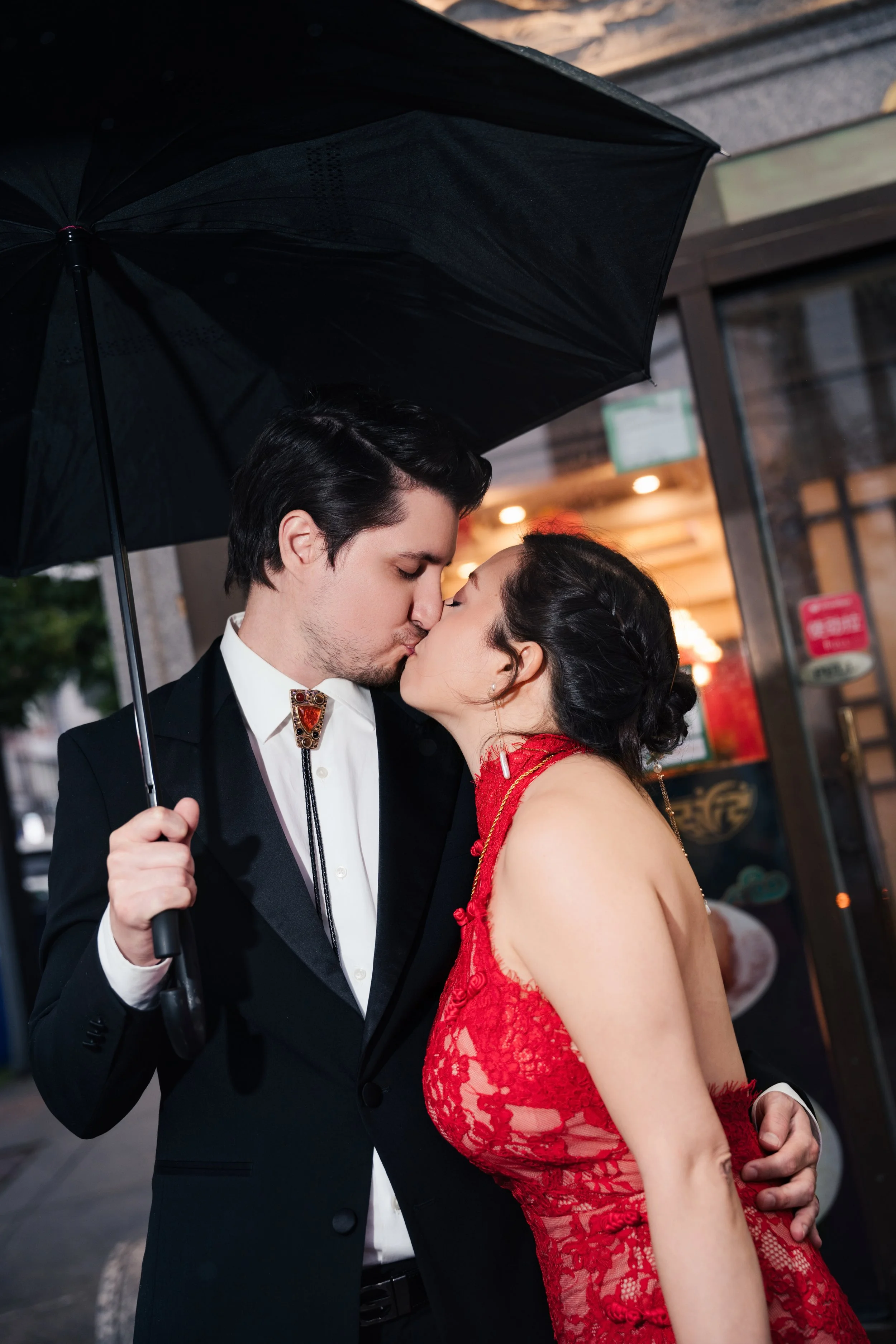 A man and woman sharing a kiss under a black umbrella on a city street at night. The man is dressed in a black tuxedo with a white shirt and decorative bolo tie, and the woman is wearing a sleeveless red lace dress.