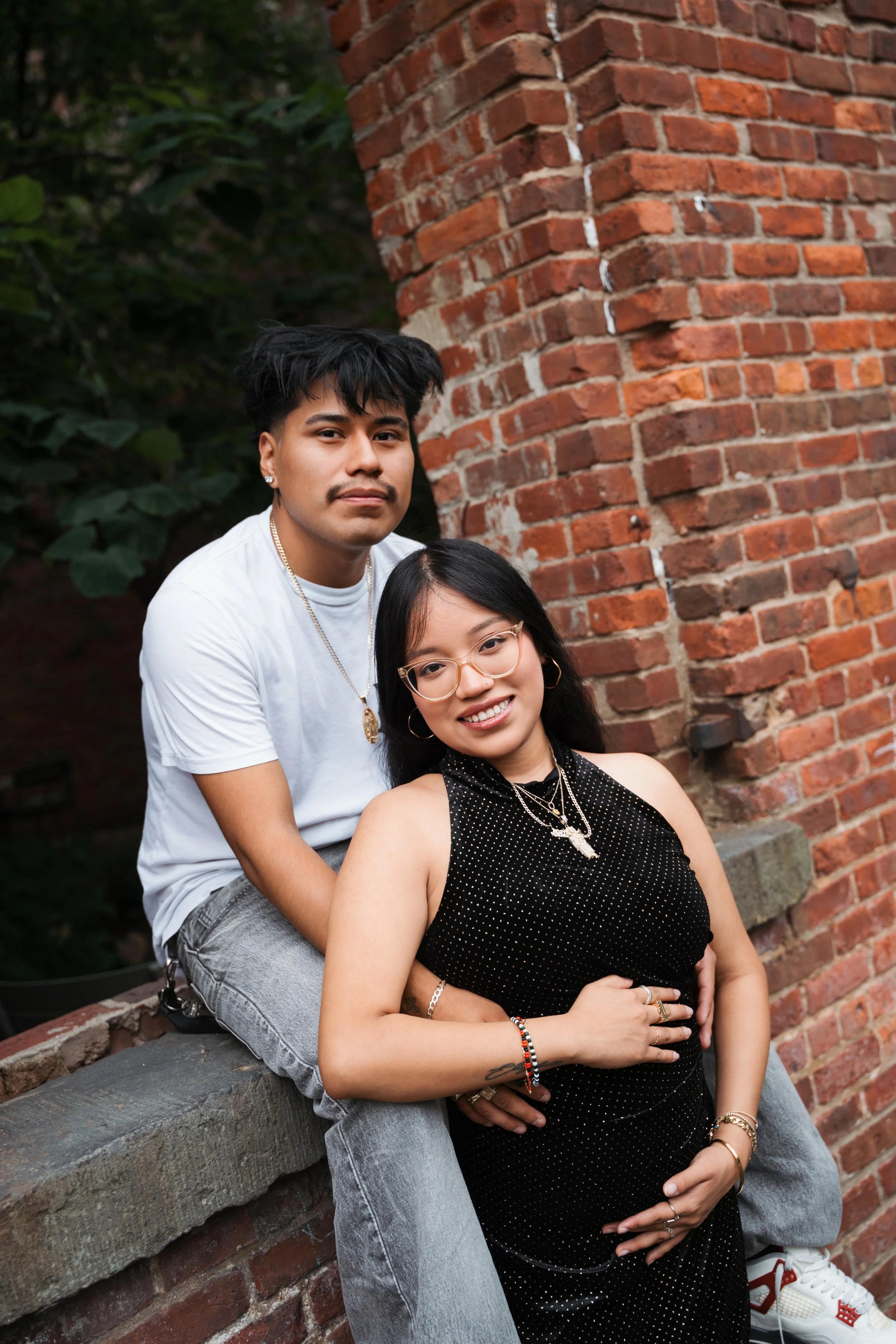 A young man and woman pose together outdoors next to a red brick wall, with greenery in the background. The woman is sitting while the man is standing behind her, both smiling.