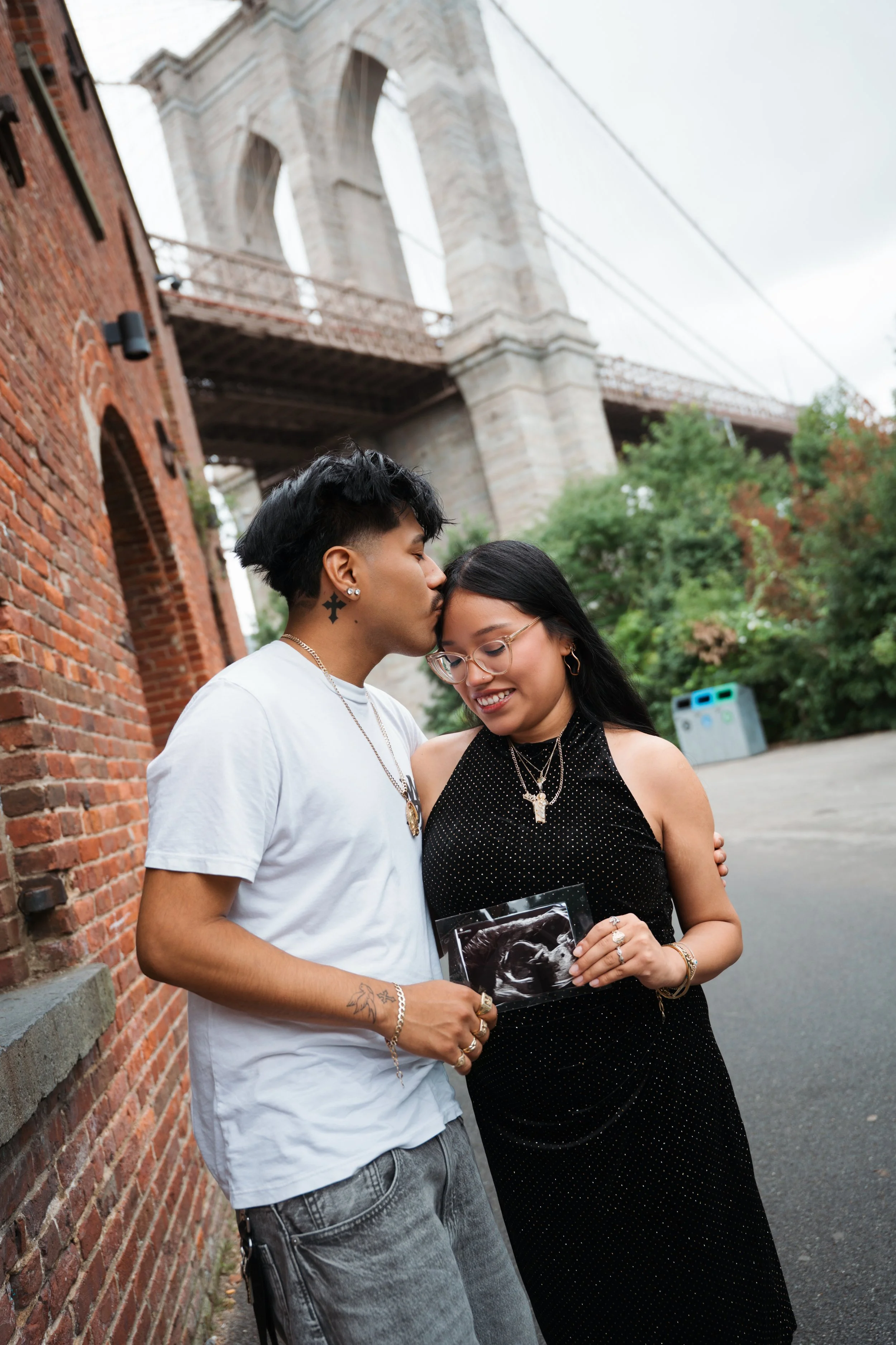 A young man and woman are standing close together on a city sidewalk, the man kissing the woman on the forehead. The woman is holding an ultrasound picture. The background features a brick building on the left and a large bridge structure overhead. It is a cloudy day with greenery and trash bins in the background.