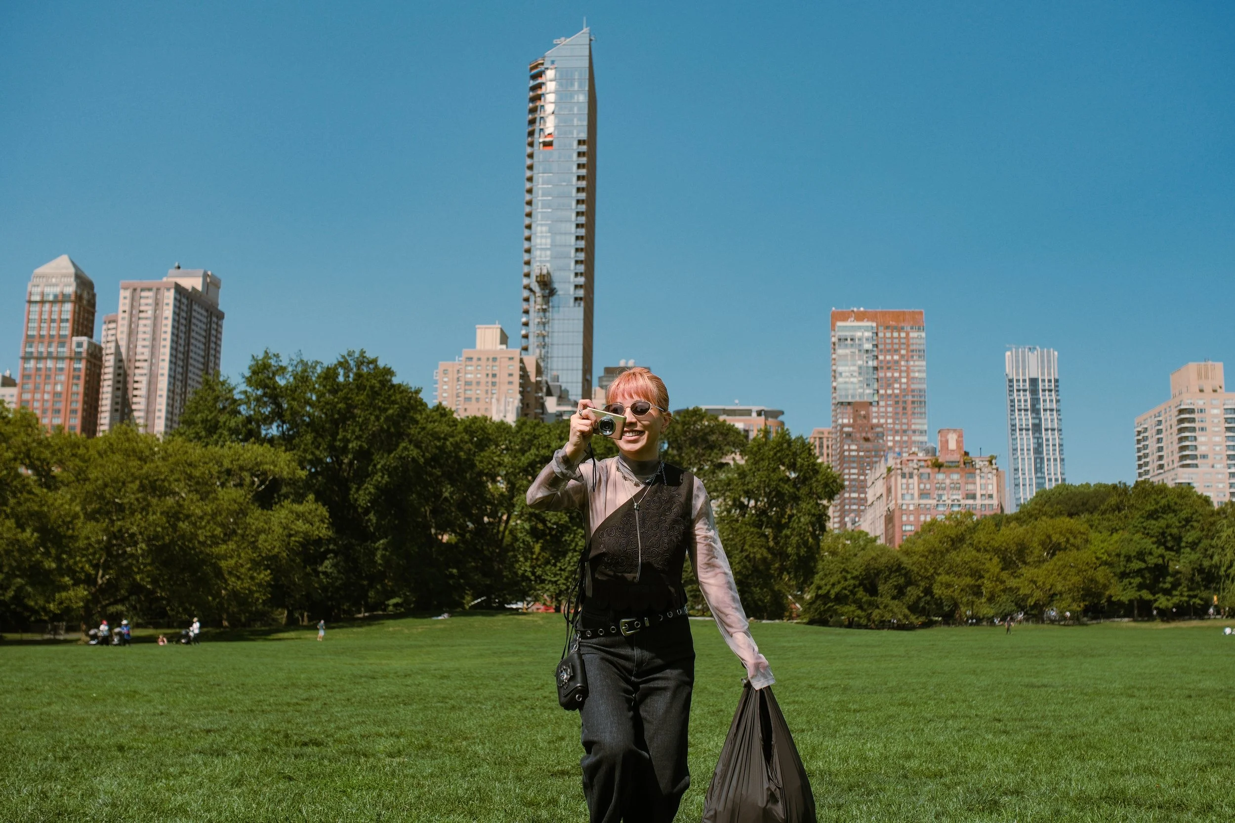 A woman with short pink hair and sunglasses smiling while taking a photo with a camera in a park with city buildings in the background.