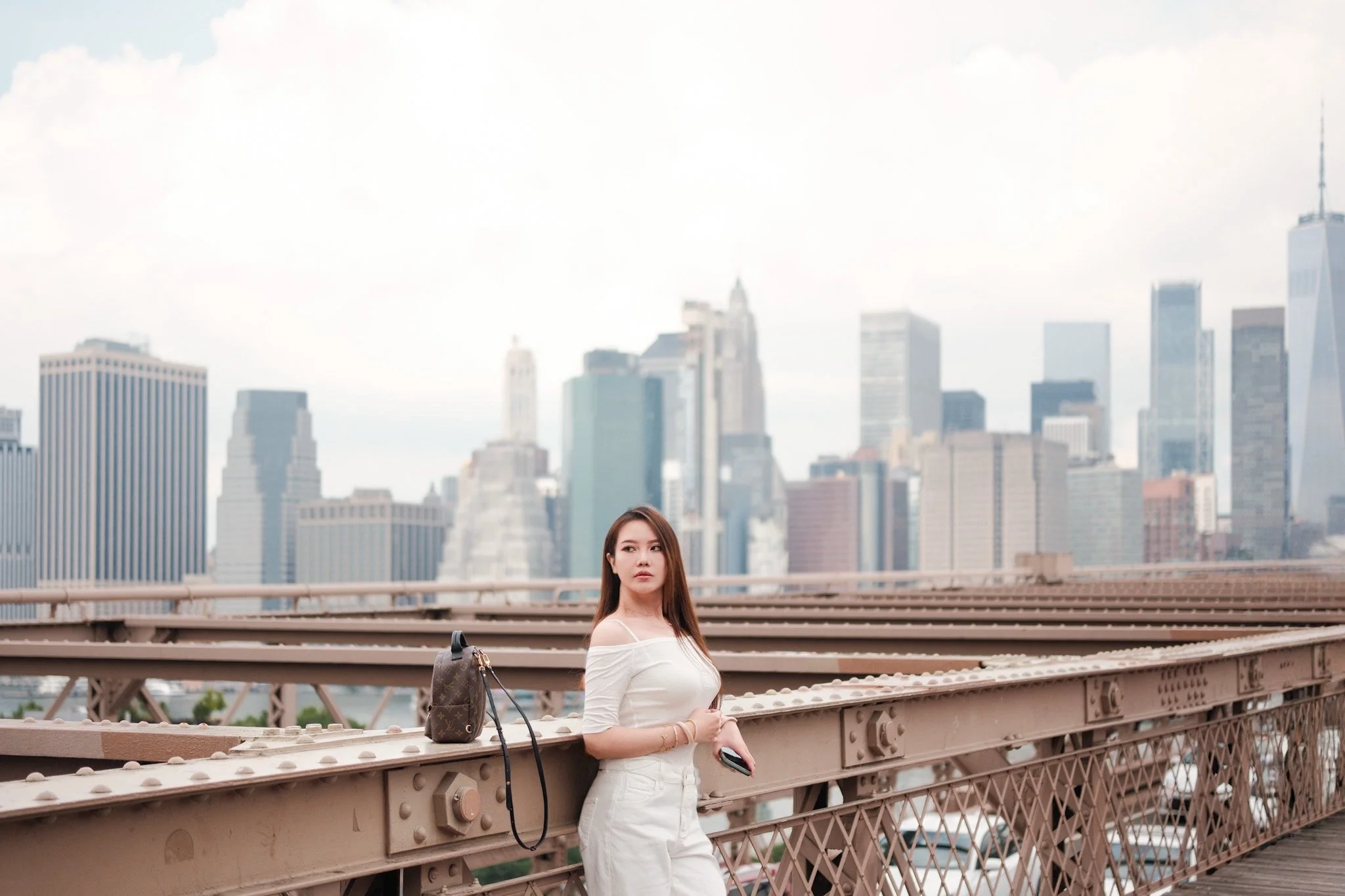 A woman with long brown hair in a white off-shoulder top and white pants holds a phone and stands on a bridge with a city skyline in the background.