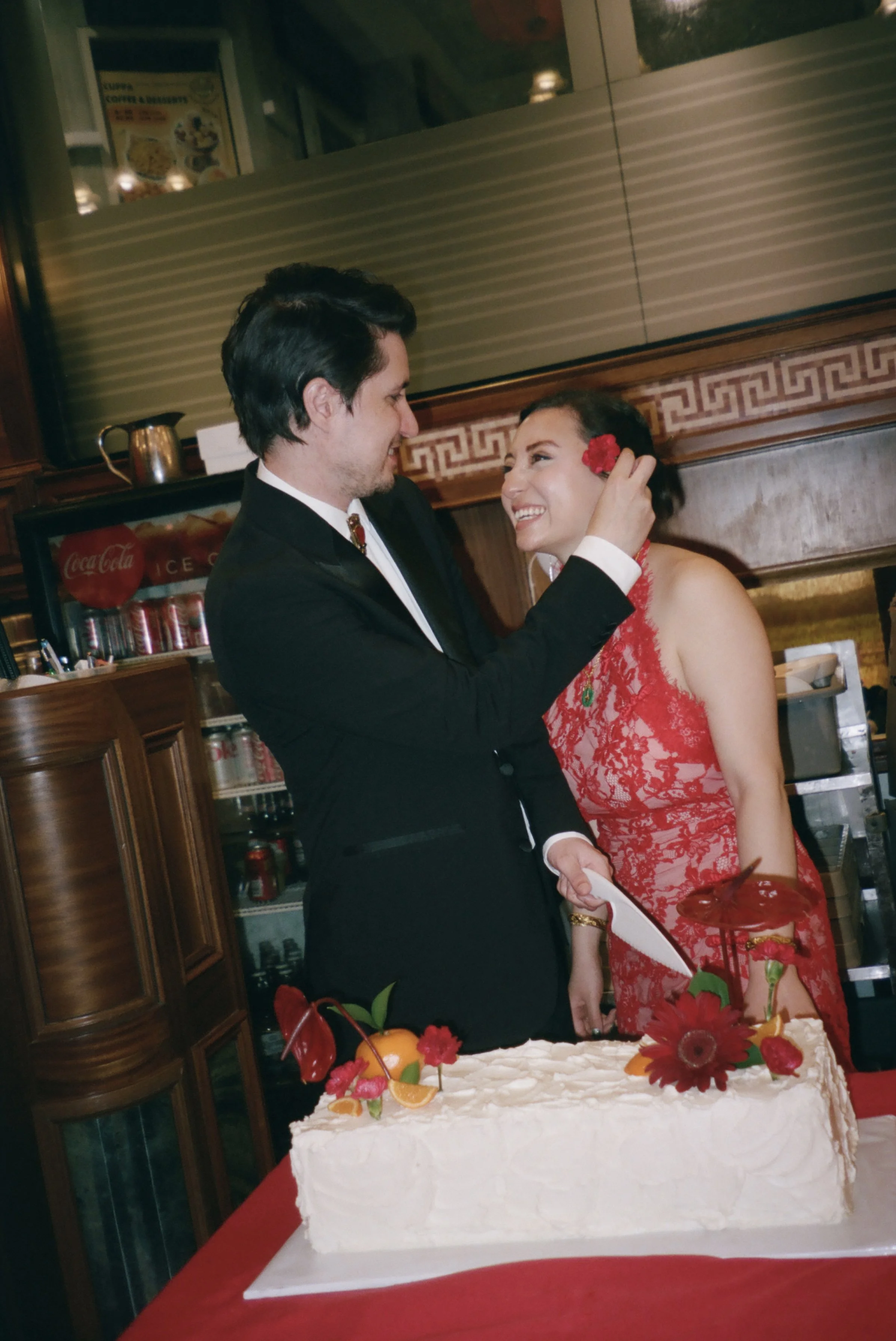 A man in a black suit and a woman in a red lace dress are smiling and looking at each other. The man is touching the woman's face, and there is a cake with flowers and fruit in front of them. The setting appears to be a restaurant or cafe.