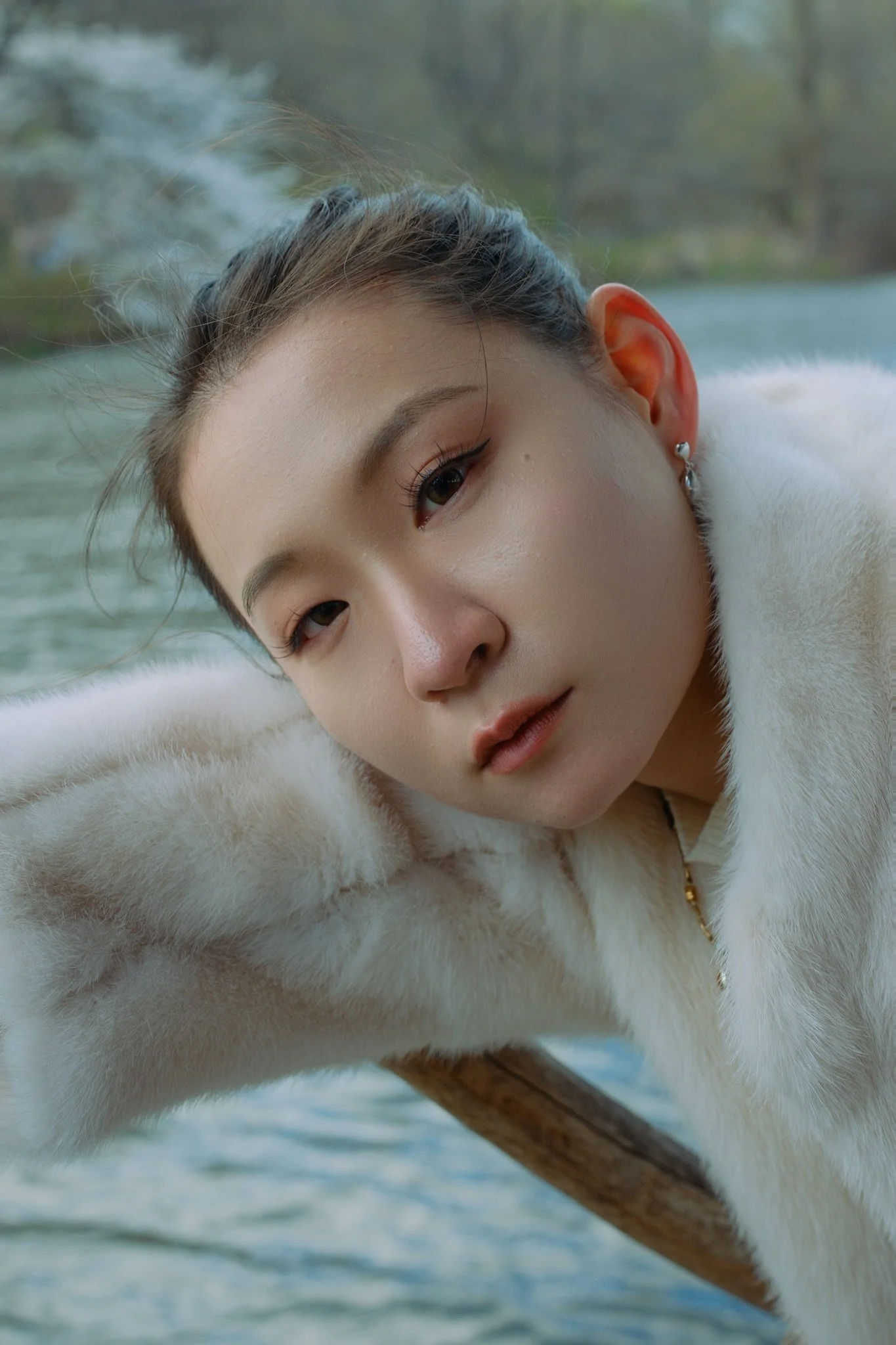 Close-up of a young woman's face with light makeup, wearing a white faux fur coat, lying outdoors near a body of water.