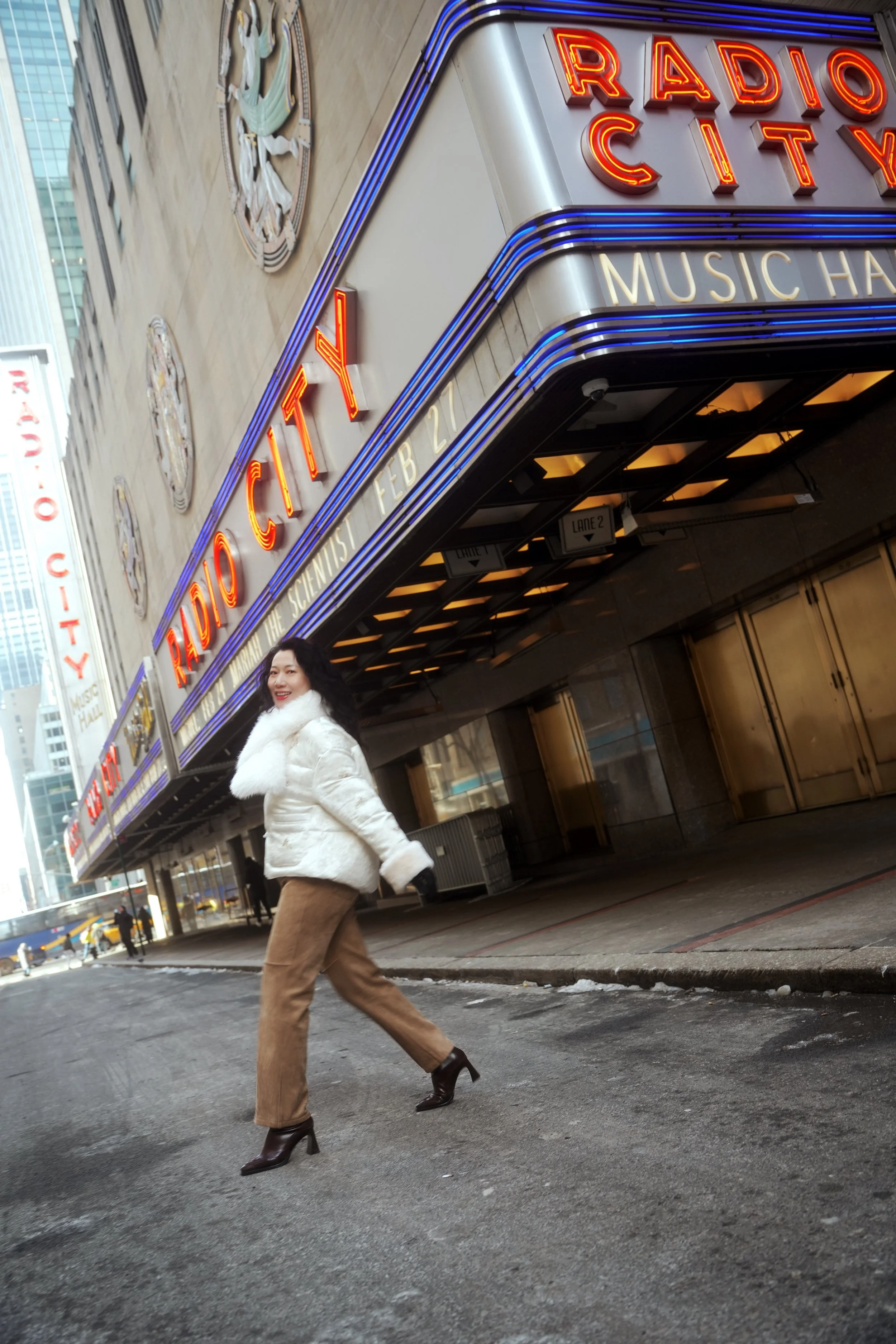 A woman wearing a white fur coat, brown pants, and high-heeled boots walking on a city sidewalk in front of a theater marquee for Radio City Music Hall with neon lights and signage.