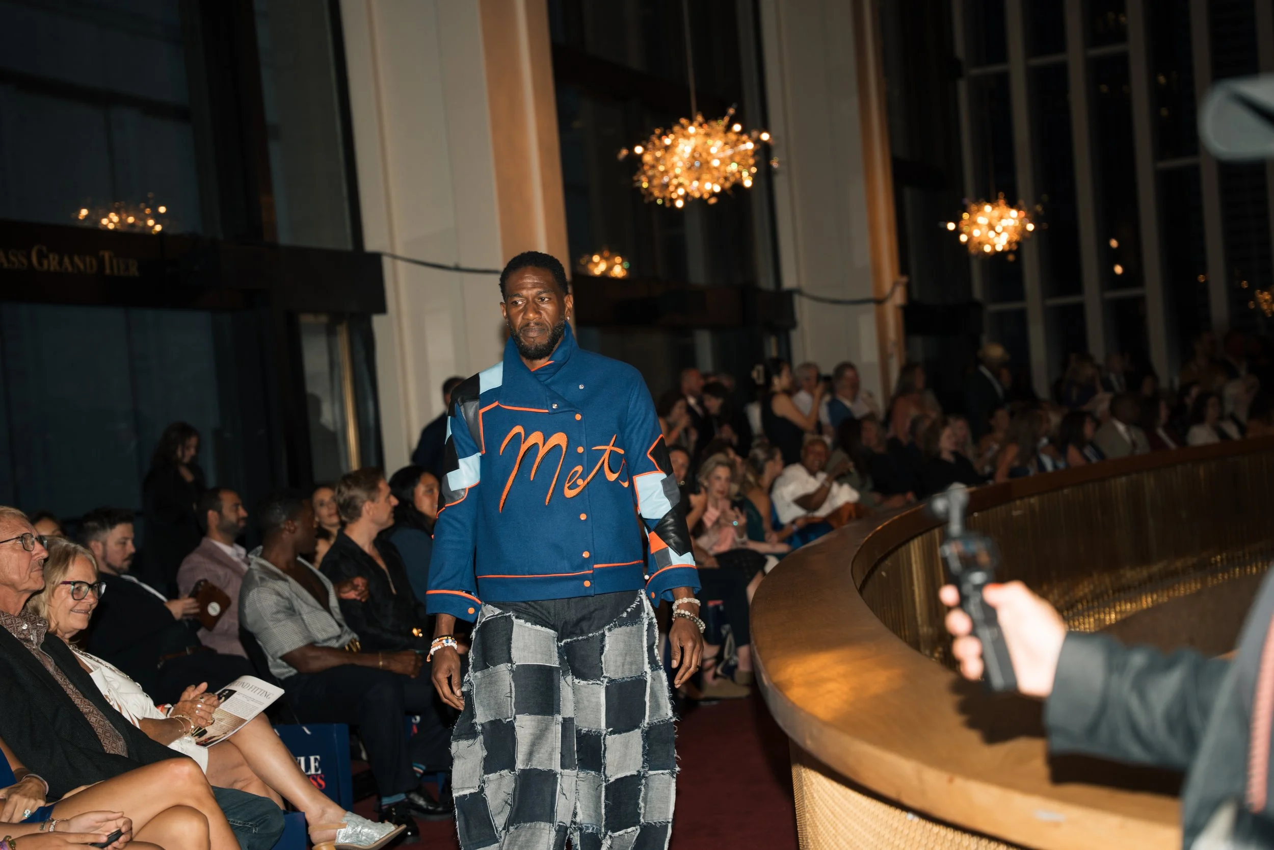 A man in a New York Mets jacket and patchwork skirt walking in a crowded auditorium with chandeliers and large windows.
