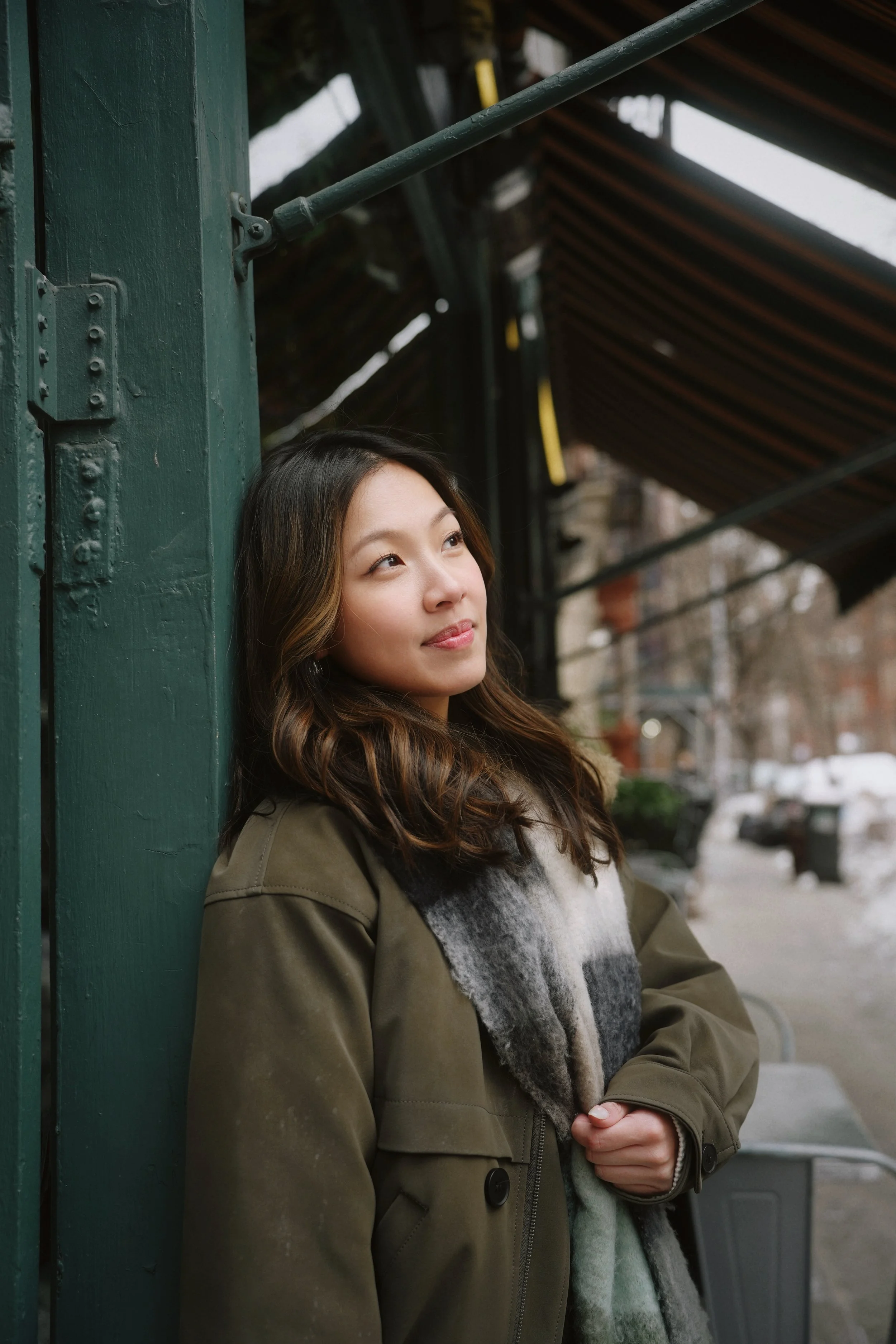 A young woman with long wavy brown hair, wearing a green jacket and a plaid sweater, is leaning against a green wooden post on a city street. She is looking slightly upwards with a calm and contemplative expression. In the background, there are build
