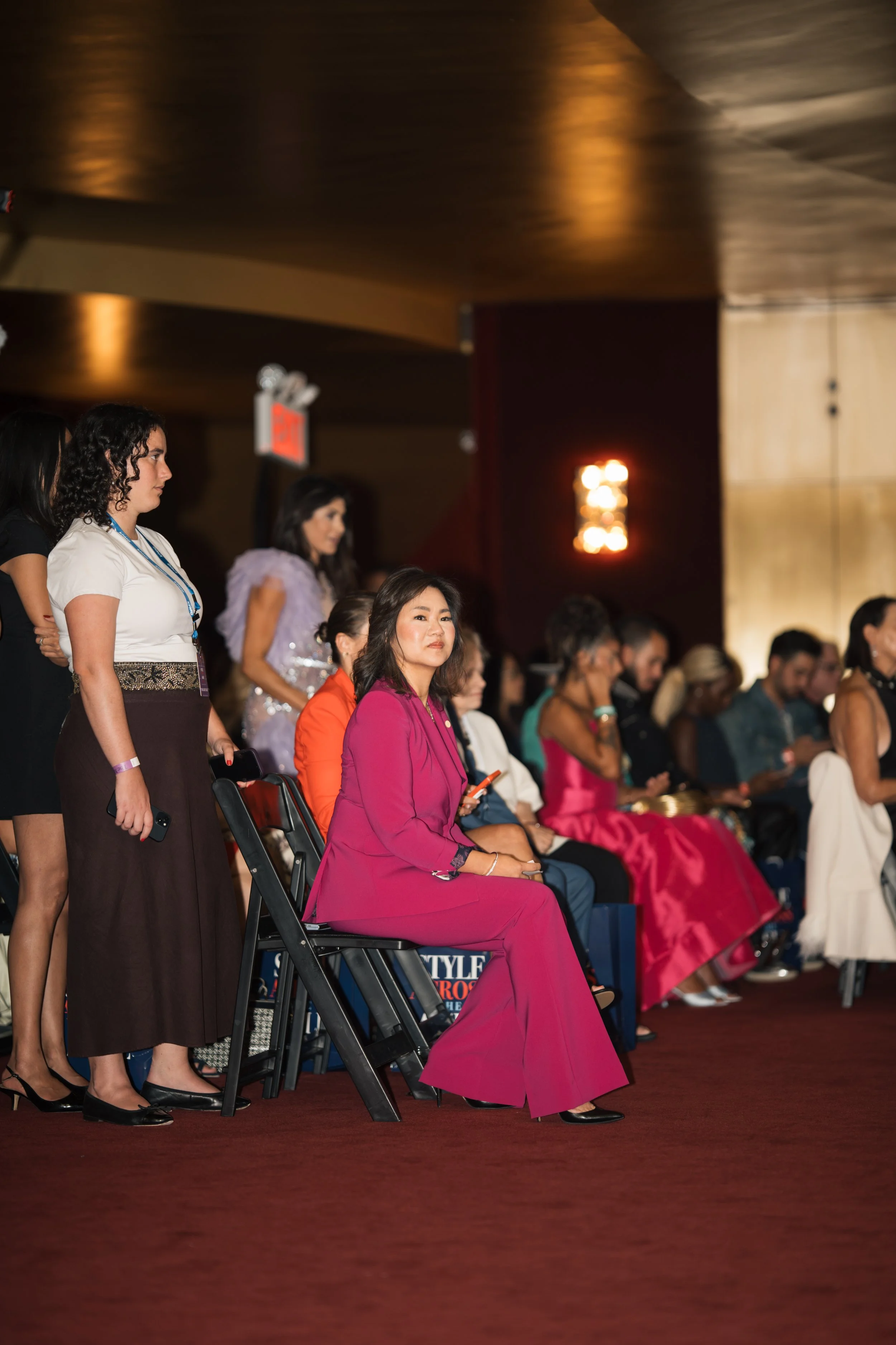 A group of people attending an event, seated in rows and standing, in a dimly lit room with wooden ceiling panels. A woman in a vibrant pink suit is seated in the foreground, while others are seen standing or seated behind her.