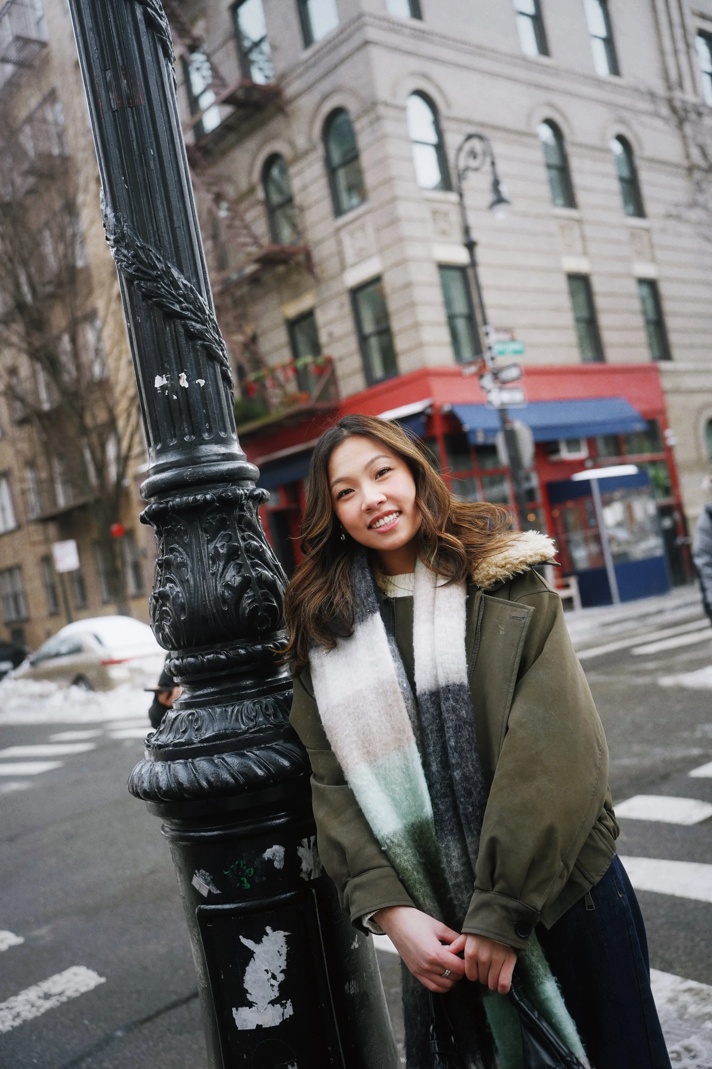 Young woman with wavy brown hair smiling, leaning against a black metal lamppost on a city street, with multi-story buildings and a crosswalk visible in the background.