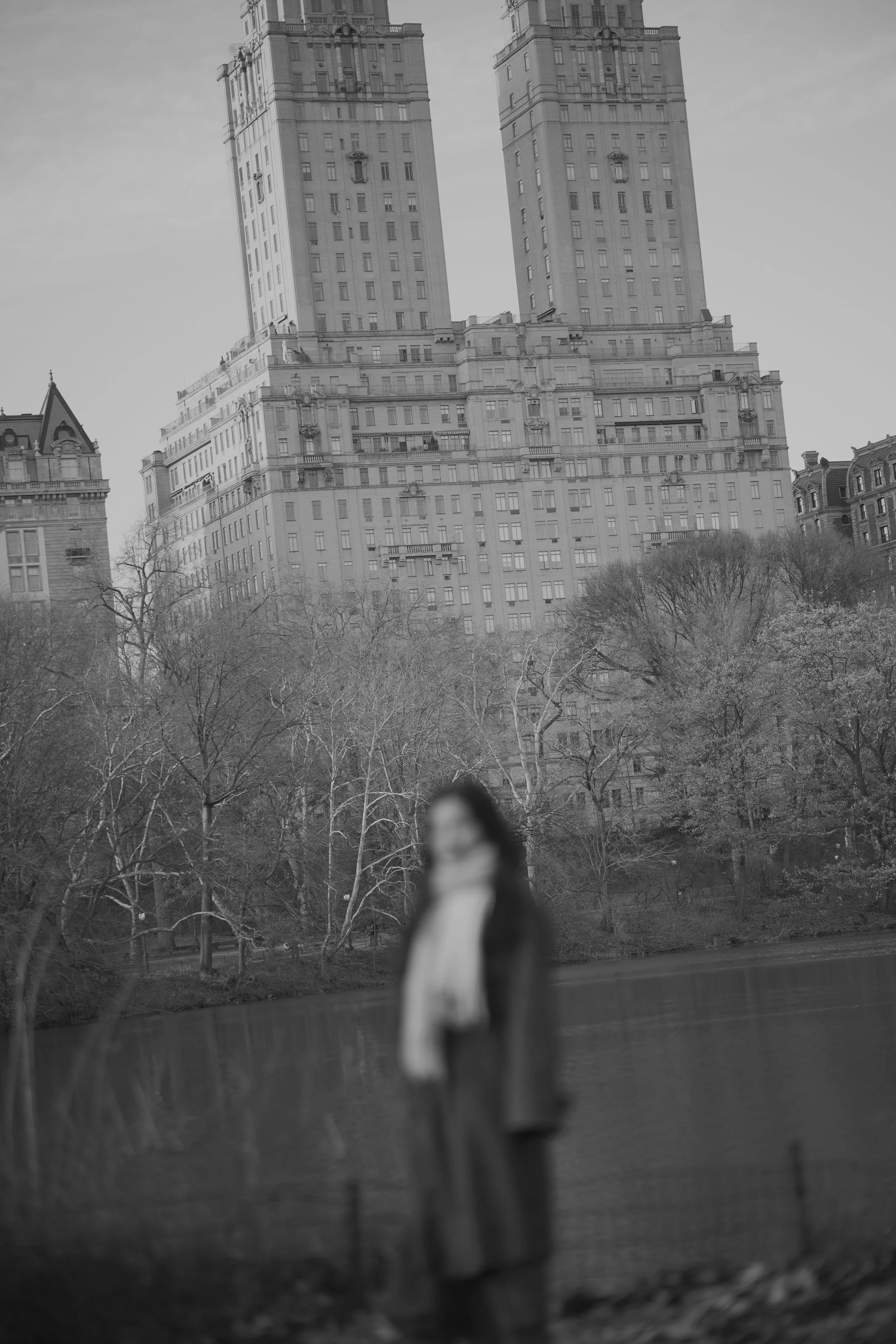 Black and white photo of a tall building with two towers, trees in the foreground, and a blurred person taking a selfie from inside a vehicle.