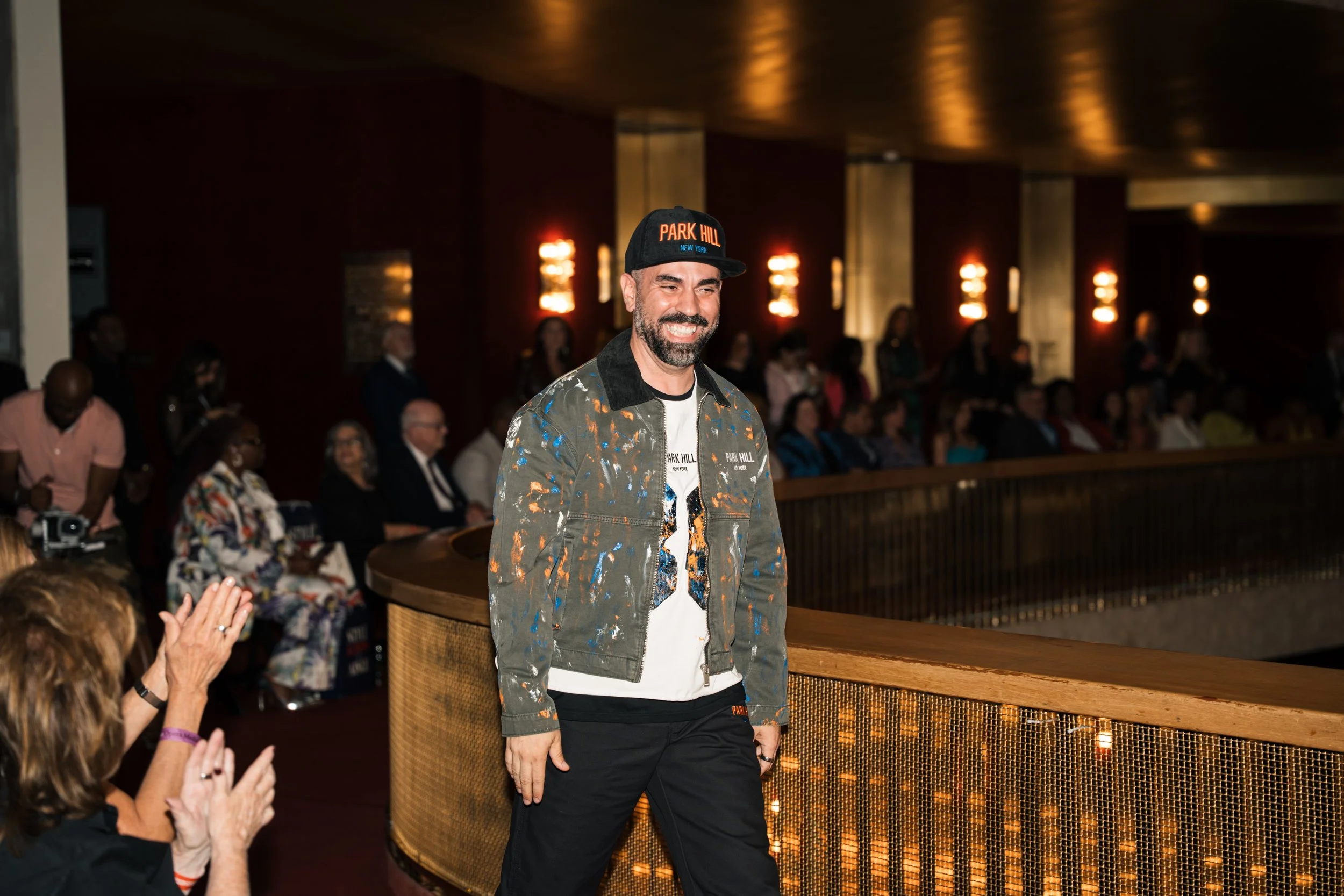 A man with a beard and short dark hair smiling and walking past an audience in a dimly lit room with warm lighting, wearing a black baseball cap that says 'PARK HILL NEW YORK' and a jacket with colorful paint splatters.