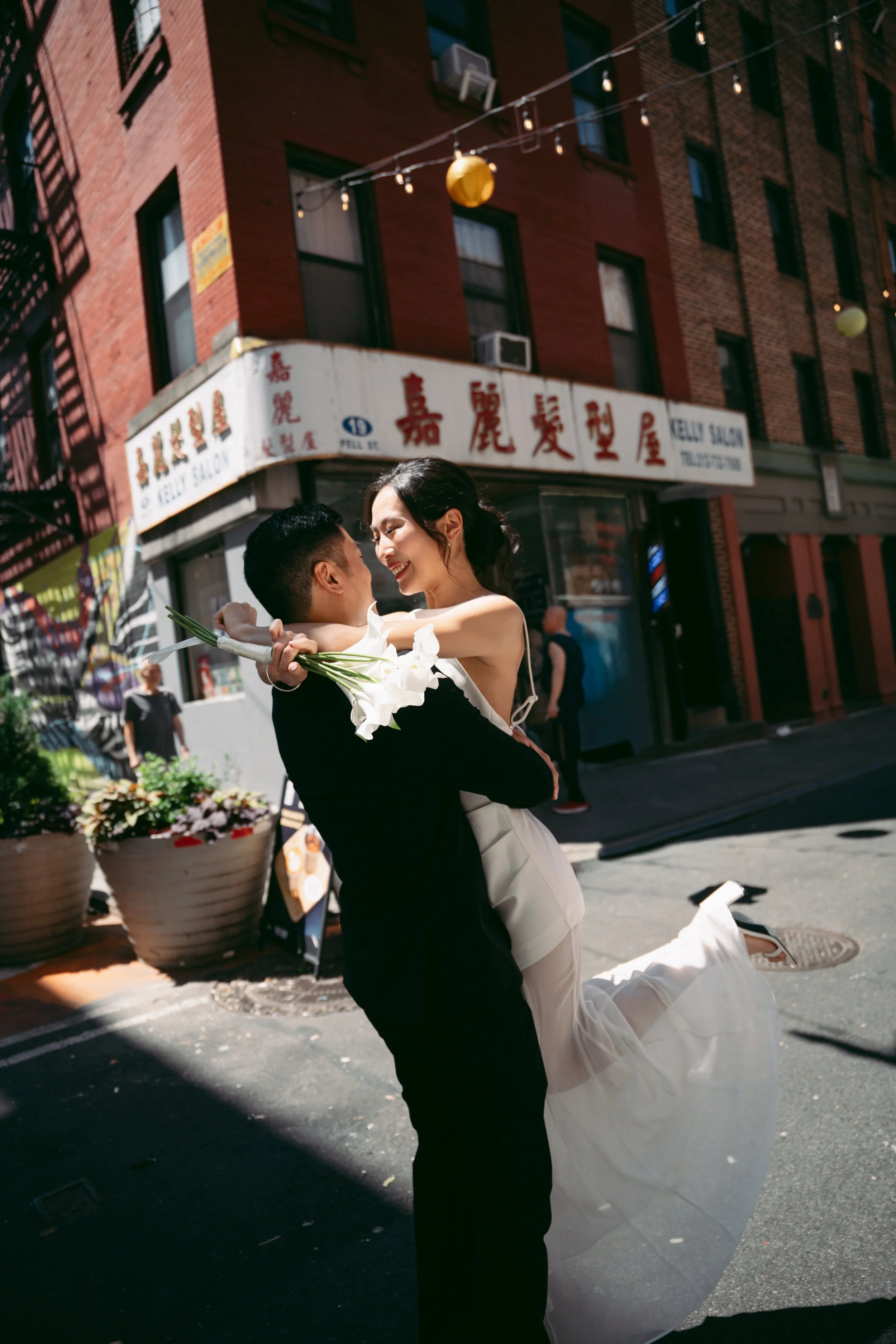 A man in formal attire is lifting and twirling a woman in a white dress with a floral bouquet in her hand, both smiling and enjoying a moment outdoors on a city street with brick buildings and hanging lanterns.