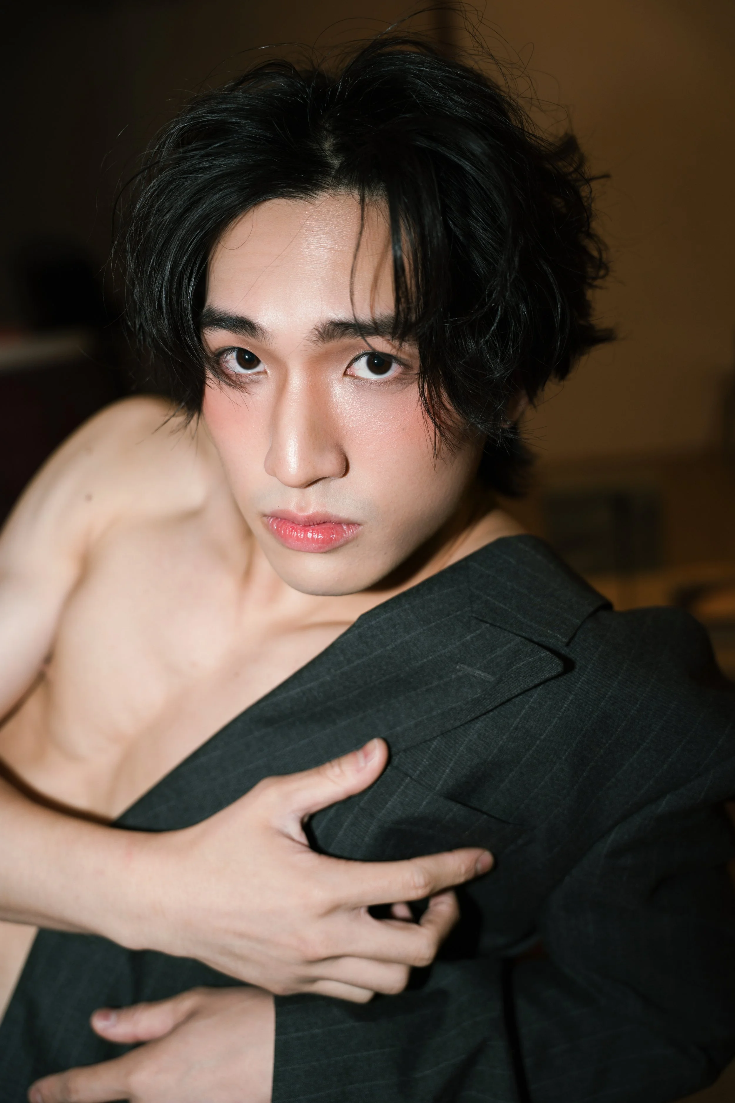 A person with black, wavy hair and fair skin wearing a dark gray pinstripe blazer, some of their shoulder and arm are exposed, and they are touching their chest with one hand.