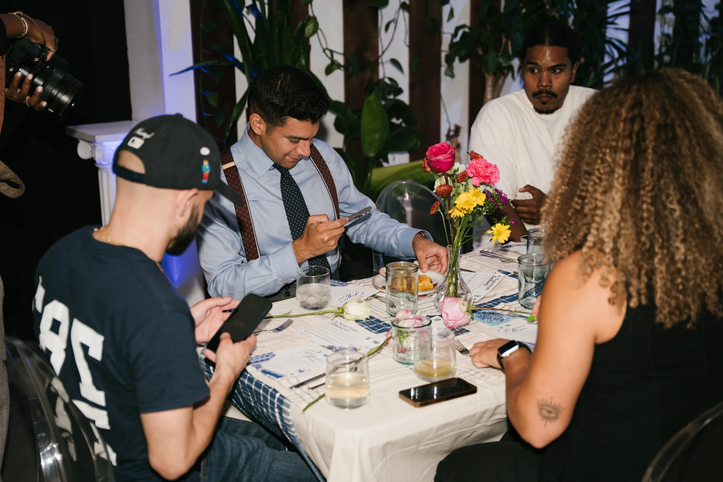 People sitting at a decorated table with flowers, glasses, and placemats, engaging in conversation during an indoor event.