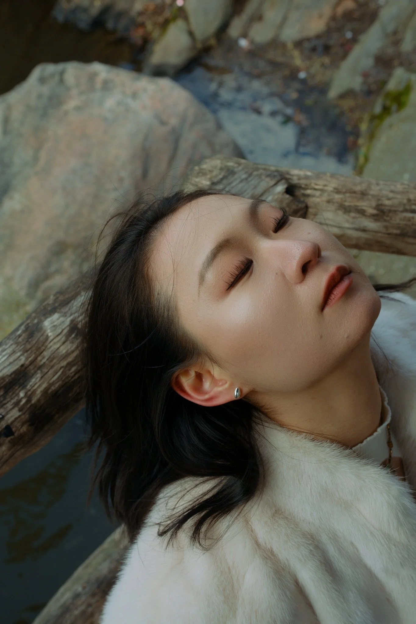 Close-up of a woman with closed eyes, lying outdoors by a wooden railing near water, with dark hair, makeup, earrings, and a white fur coat or scarf.
