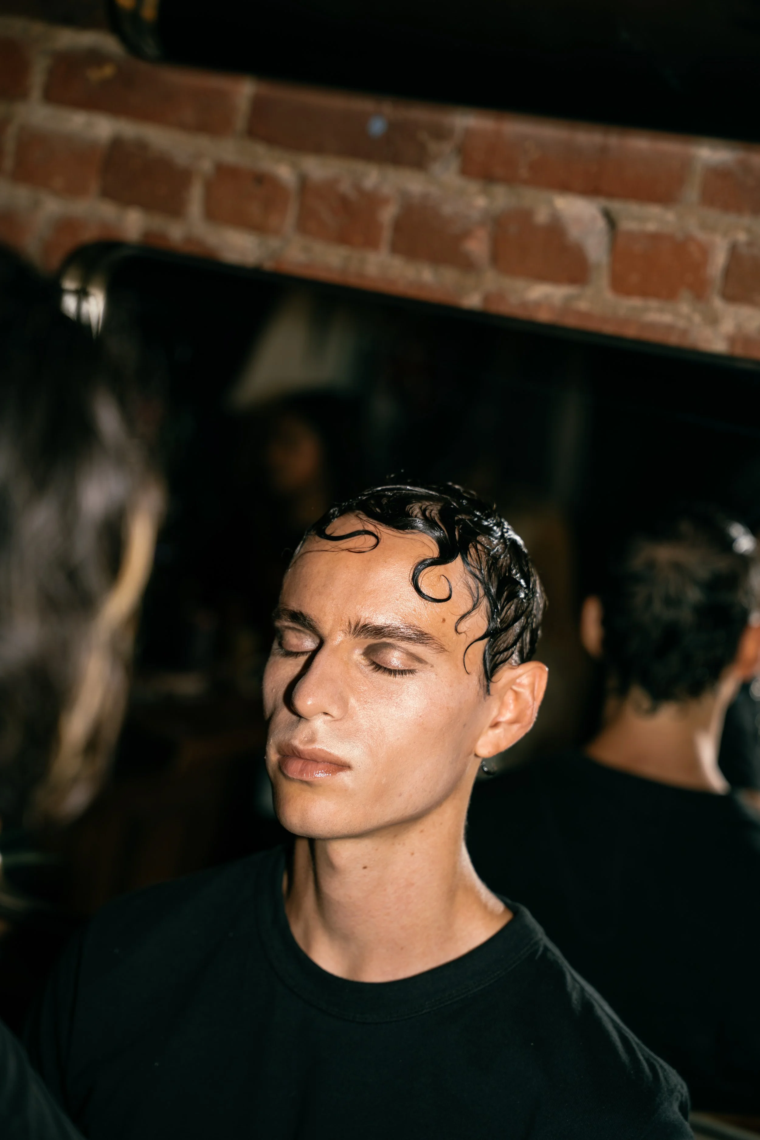 Close-up of a young man with wet, styled hair and a black shirt, with a brick wall background and blurred figures around him.