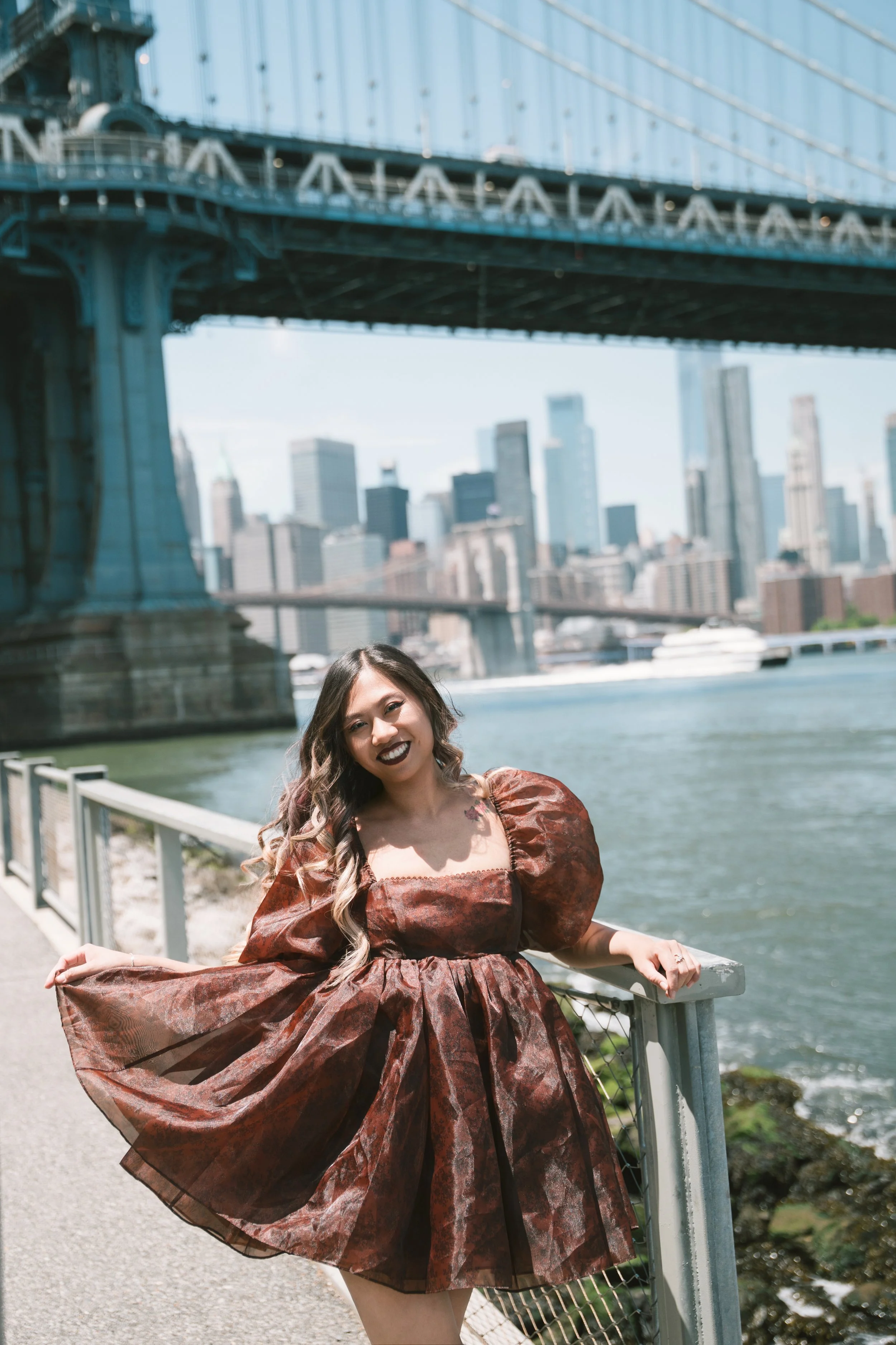 A woman wearing a large, patterned, brown dress with puffy sleeves standing by a railing next to a river with a city skyline and a bridge in the background.