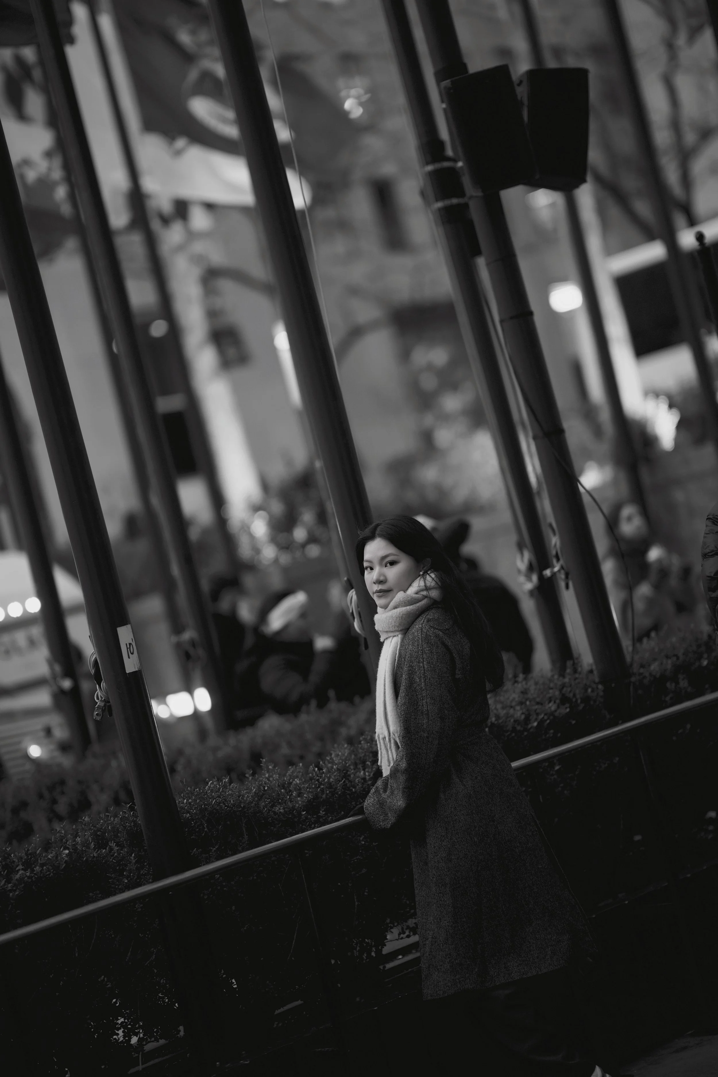 A woman with long dark hair and a light-colored scarf stands outdoors near a reflective glass building, with trees and parked cars in the background, in black and white.