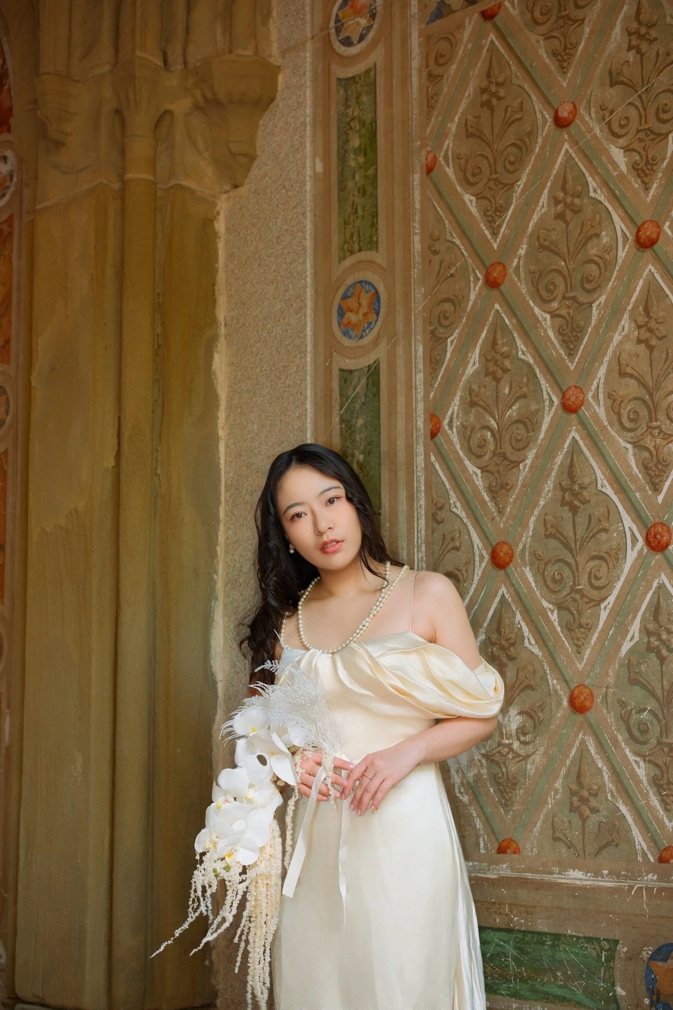 Young woman in a light-colored, elegant dress holding a bouquet of white flowers, standing by an ornate wall with intricate patterns and a large wooden column in the background.