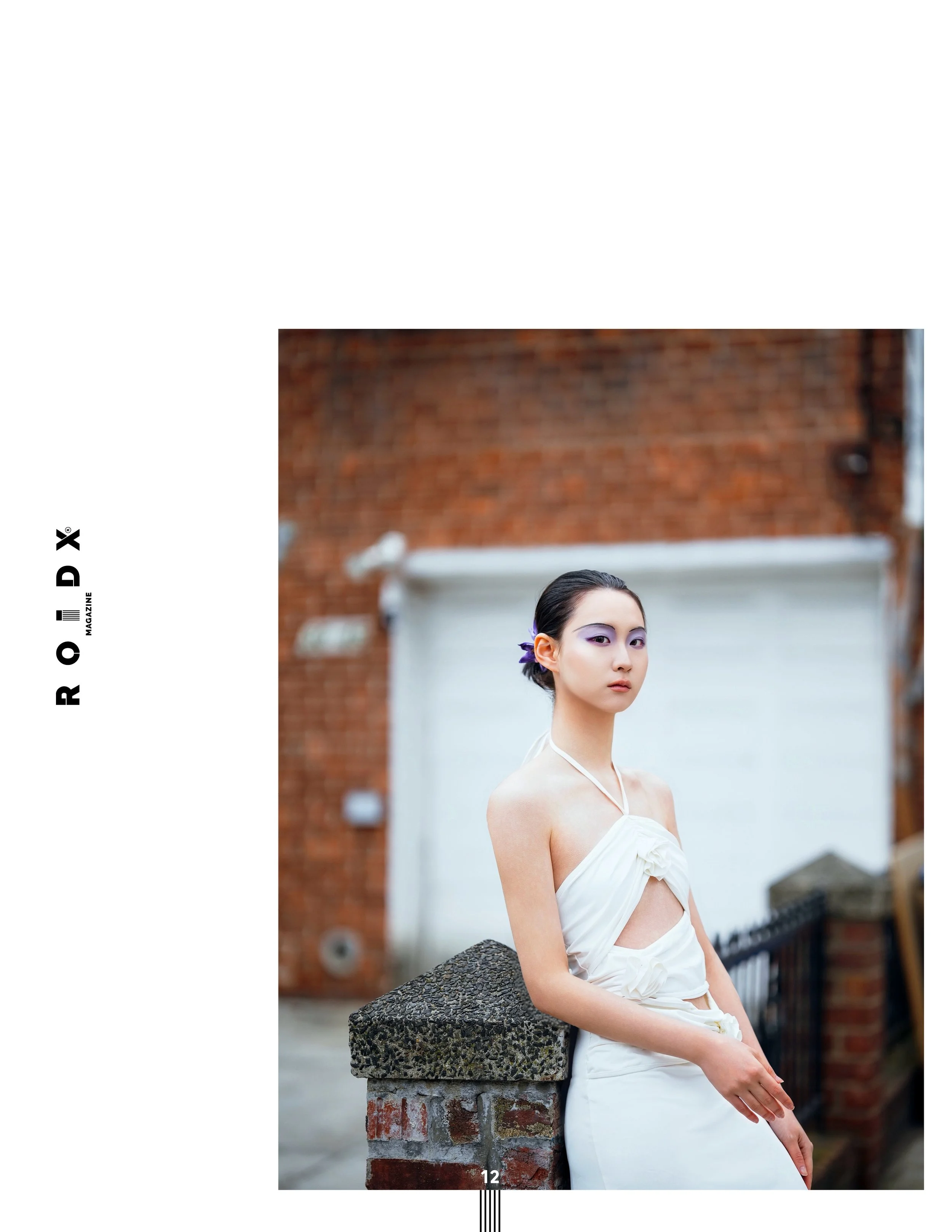 A young woman with dark hair styled back, wearing purple eye makeup and a white sleeveless dress with a knotted front, sitting on a stone bench with her arm extended, in an outdoor setting with brick building and black fence in the background.