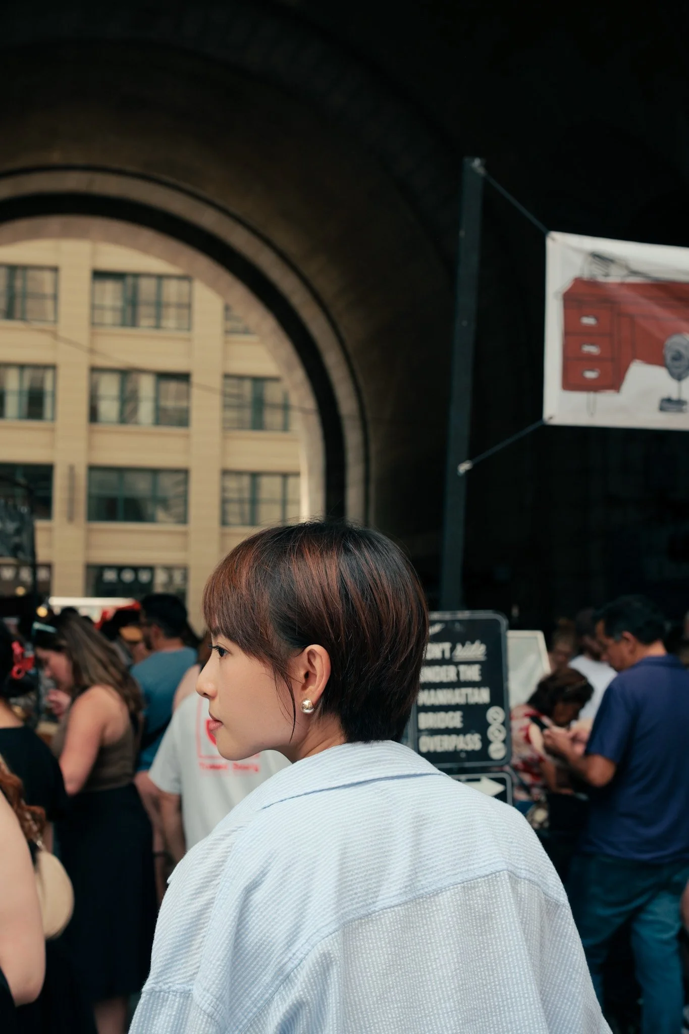 A woman with short hair, pearl earrings, and a light-colored shirt in a crowded outdoor setting with people, signboards, and a building in the background.