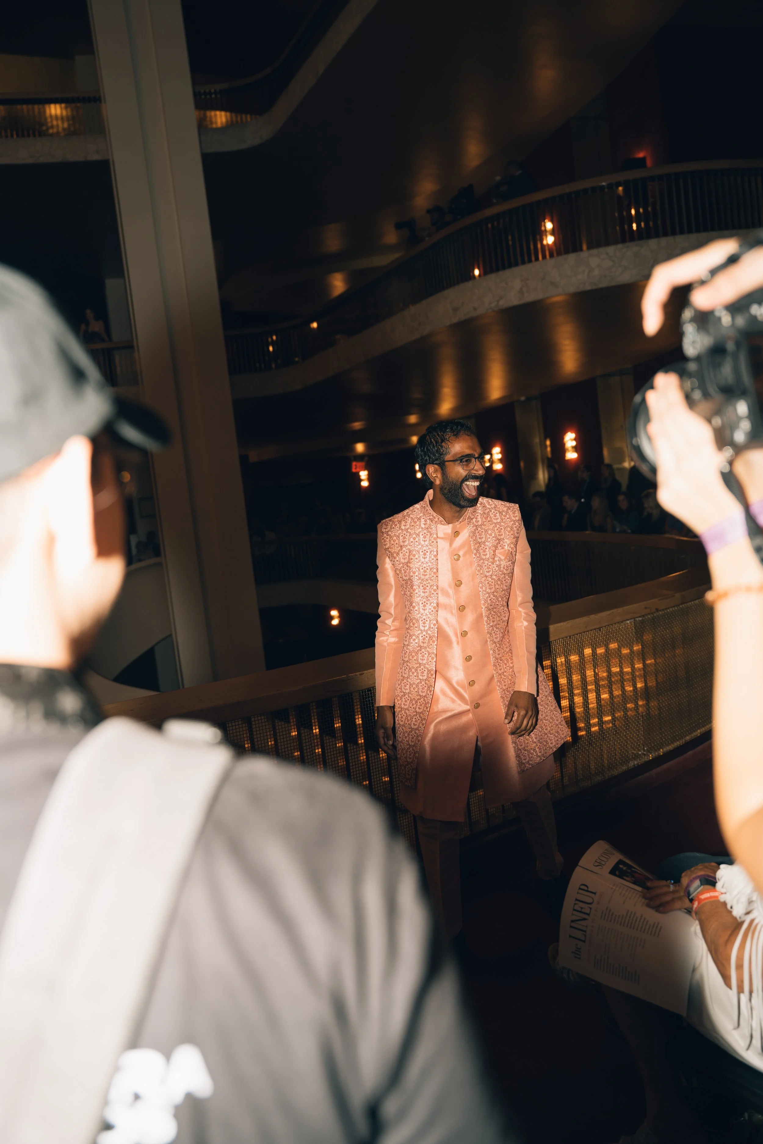 Man dressed in traditional Indian attire, smiling and standing in an indoor event space while others, including photographers, are capturing the moment.