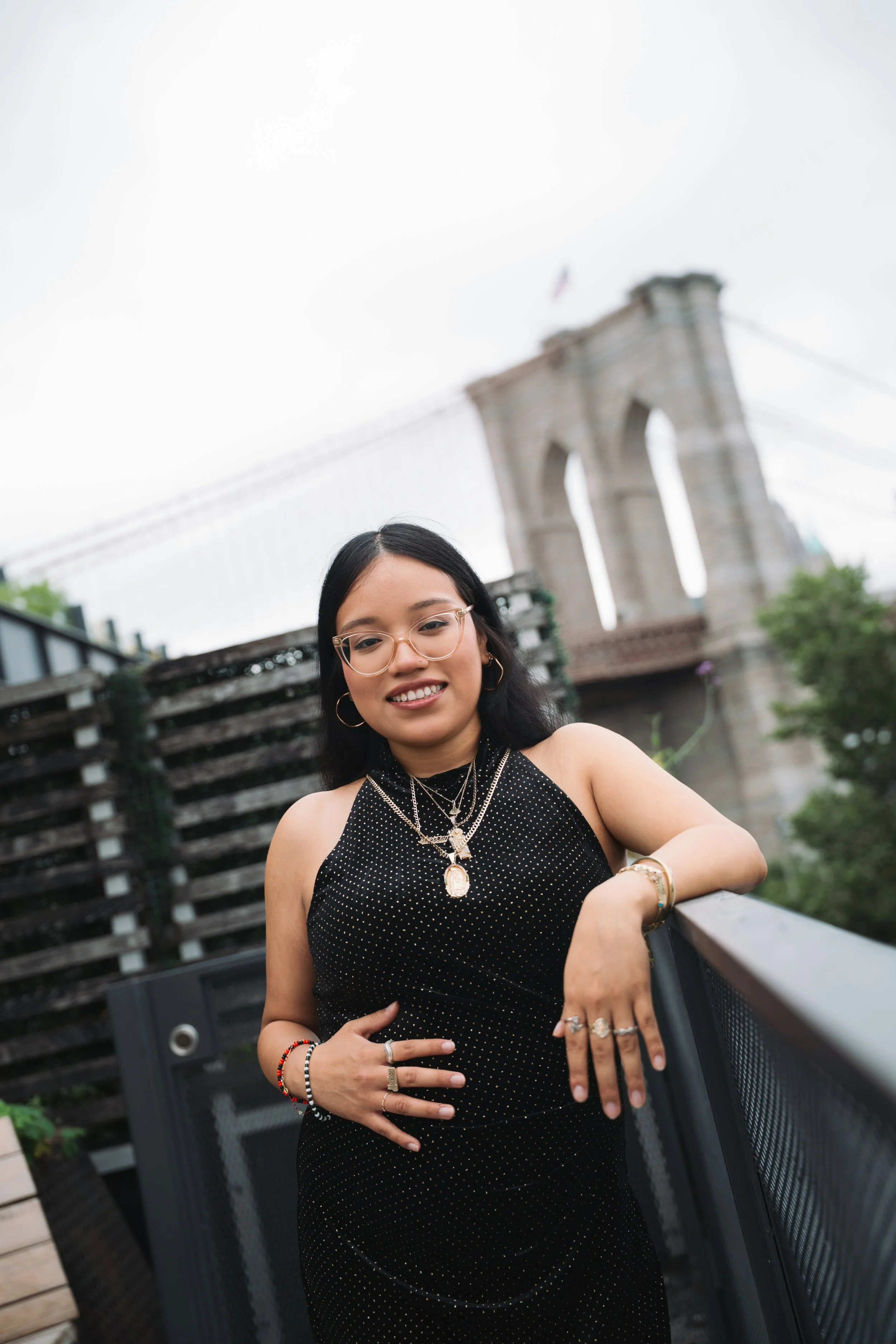 A woman with long dark hair and glasses wearing a black dress with white polka dots, jewelry including necklaces and rings, standing outdoors near the Brooklyn Bridge in New York City, smiling and resting her hand on a railing.