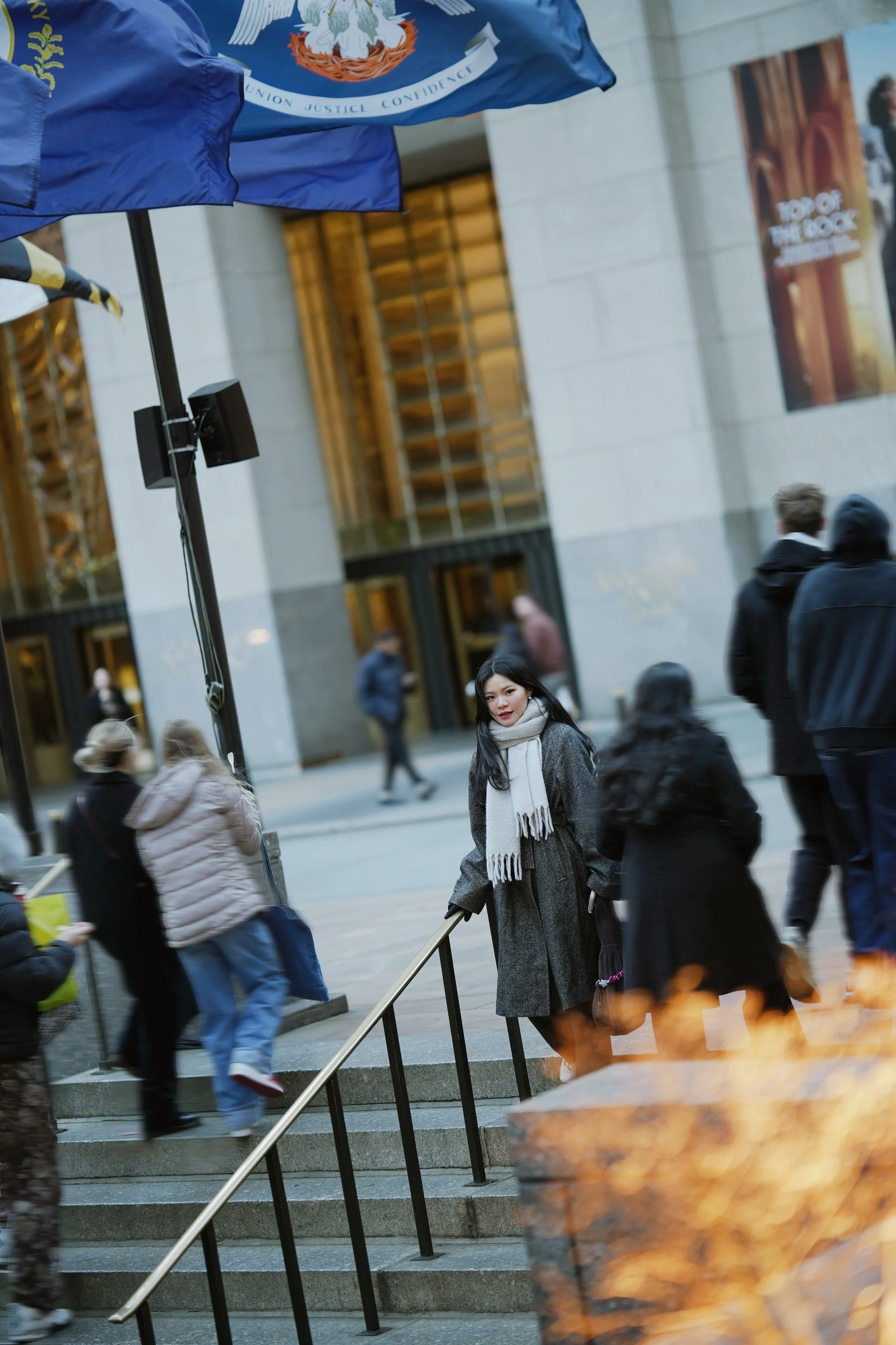 A woman with dark hair, wearing a gray coat and white scarf, standing among pedestrians outside a building with wooden accents. There are flags, including one with a seal, and photo posters on the building wall. The scene appears to be in an urban ar