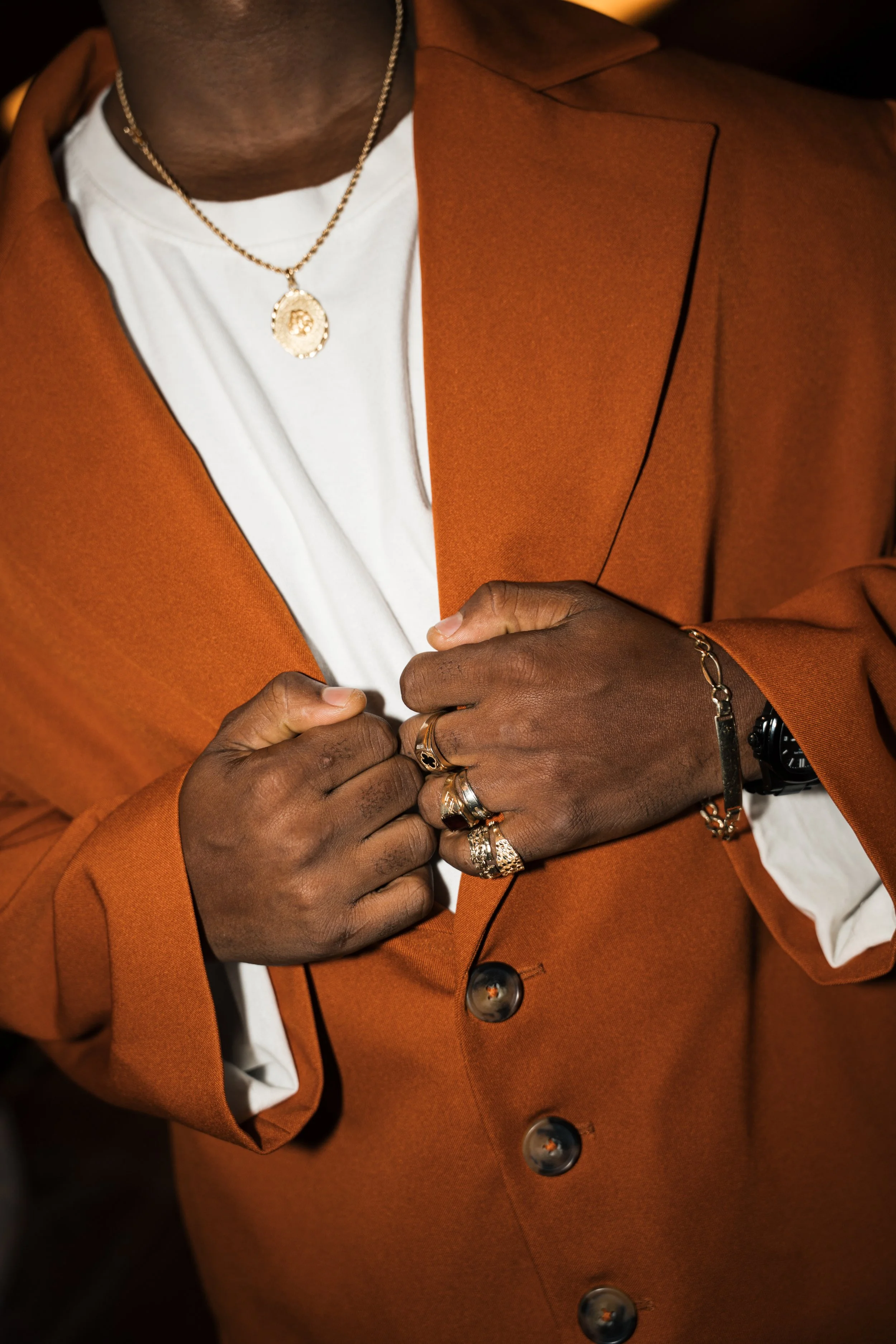 Close-up of a person wearing a white shirt, an orange blazer, gold jewelry, and a black watch, adjusting the blazer with their hands.