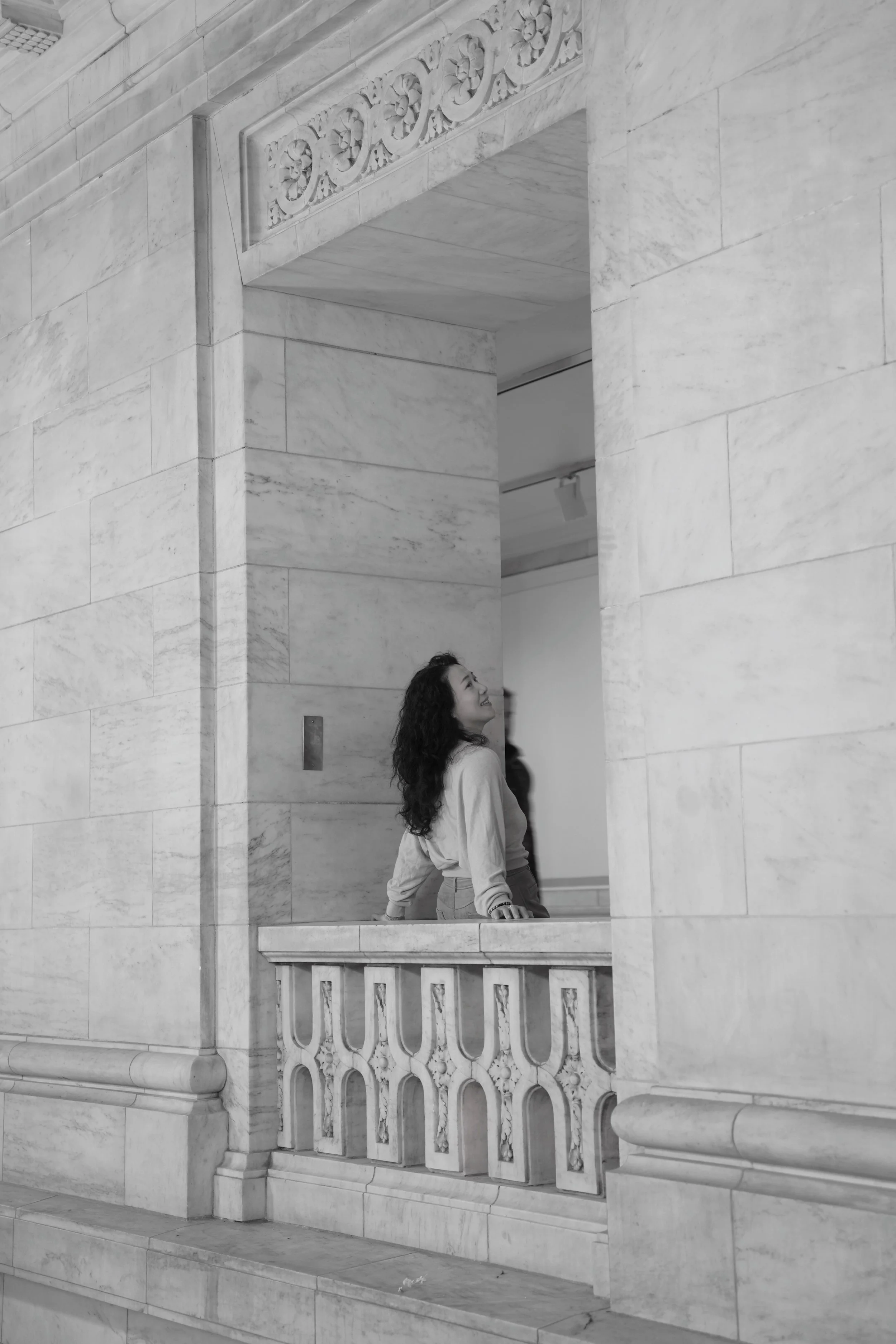 A woman with long curly hair leaning on a balcony railing inside a marble building, looking up and smiling.