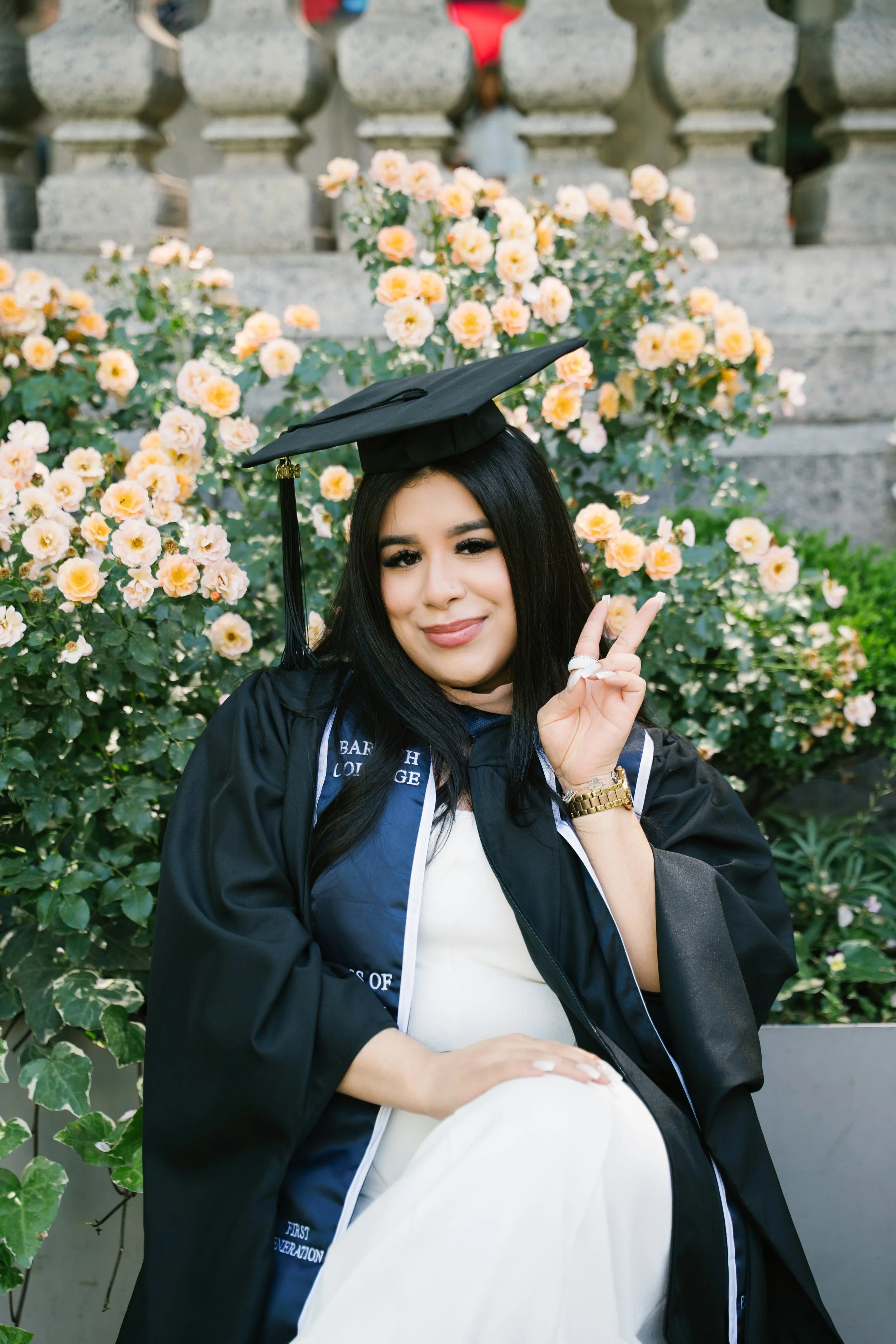 Young woman in graduation cap and gown making a peace sign while sitting outdoors near blooming bushes.