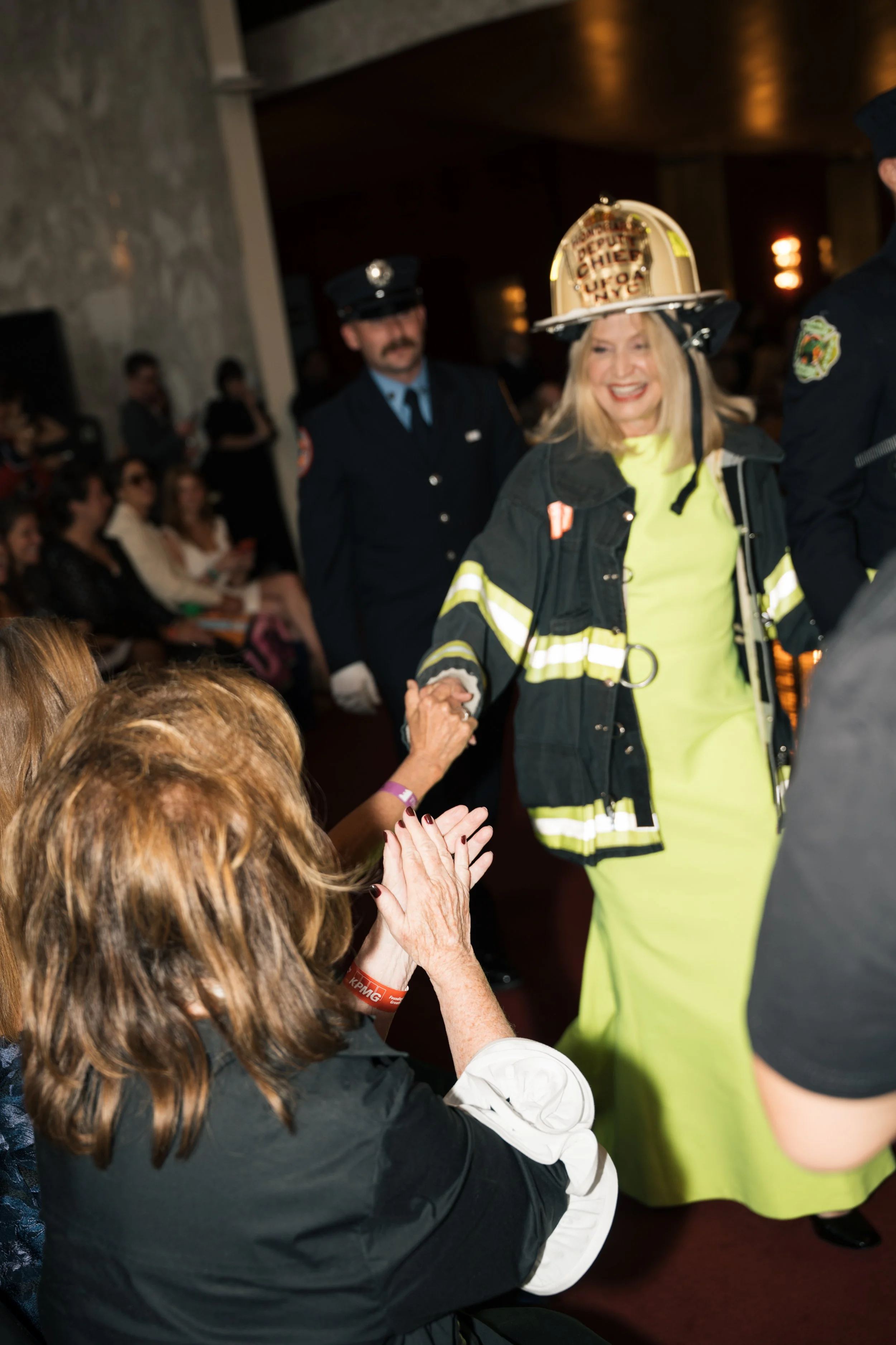 A woman in a firefighter's uniform holding hands and smiling with an audience of seated women and people dressed in firefighter uniforms at an indoor event.