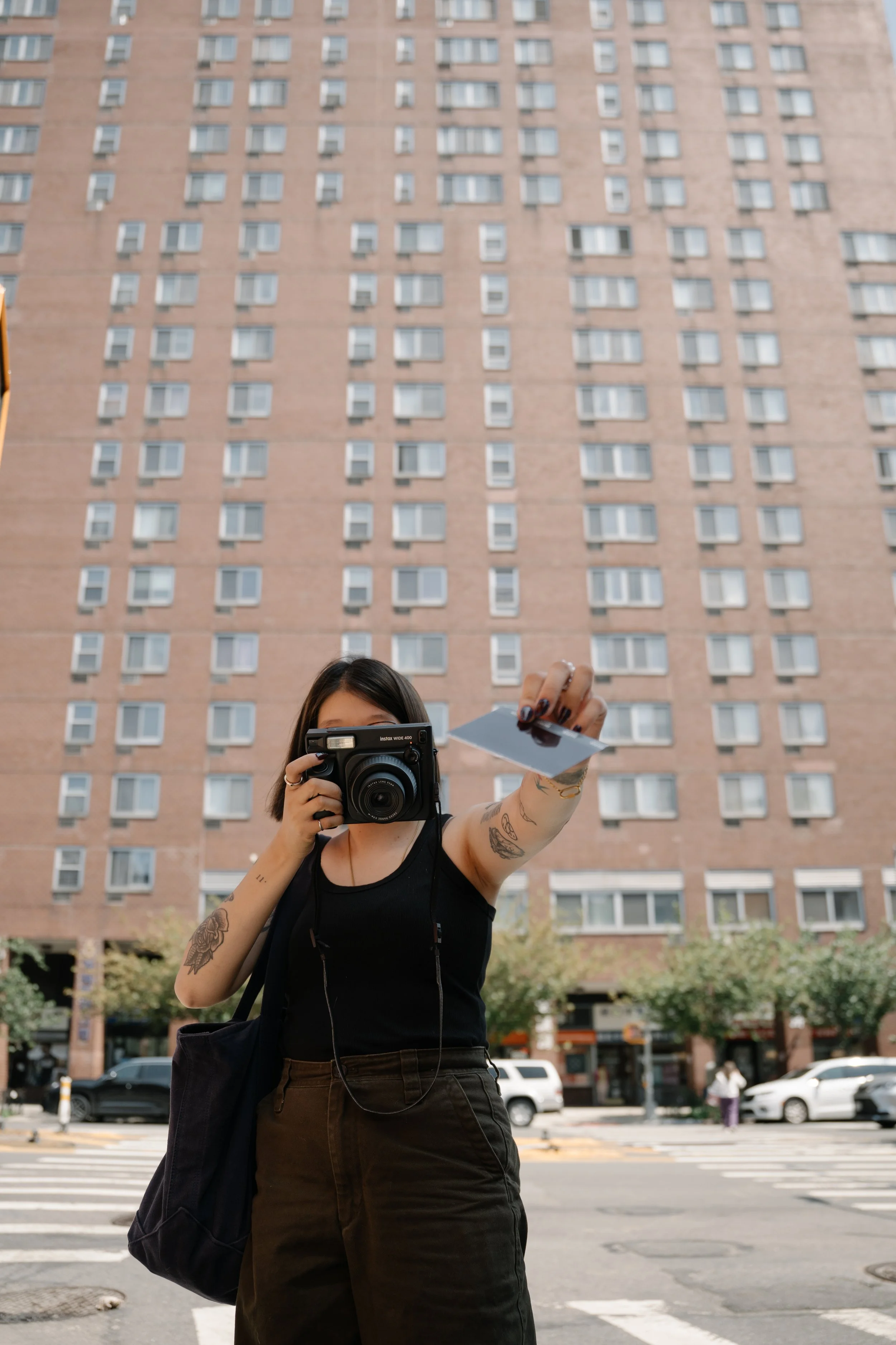 A woman with tattoos on her arms, wearing a black sleeveless top, taking a photo with a camera in an urban street setting with a tall brick apartment building and cars in the background.