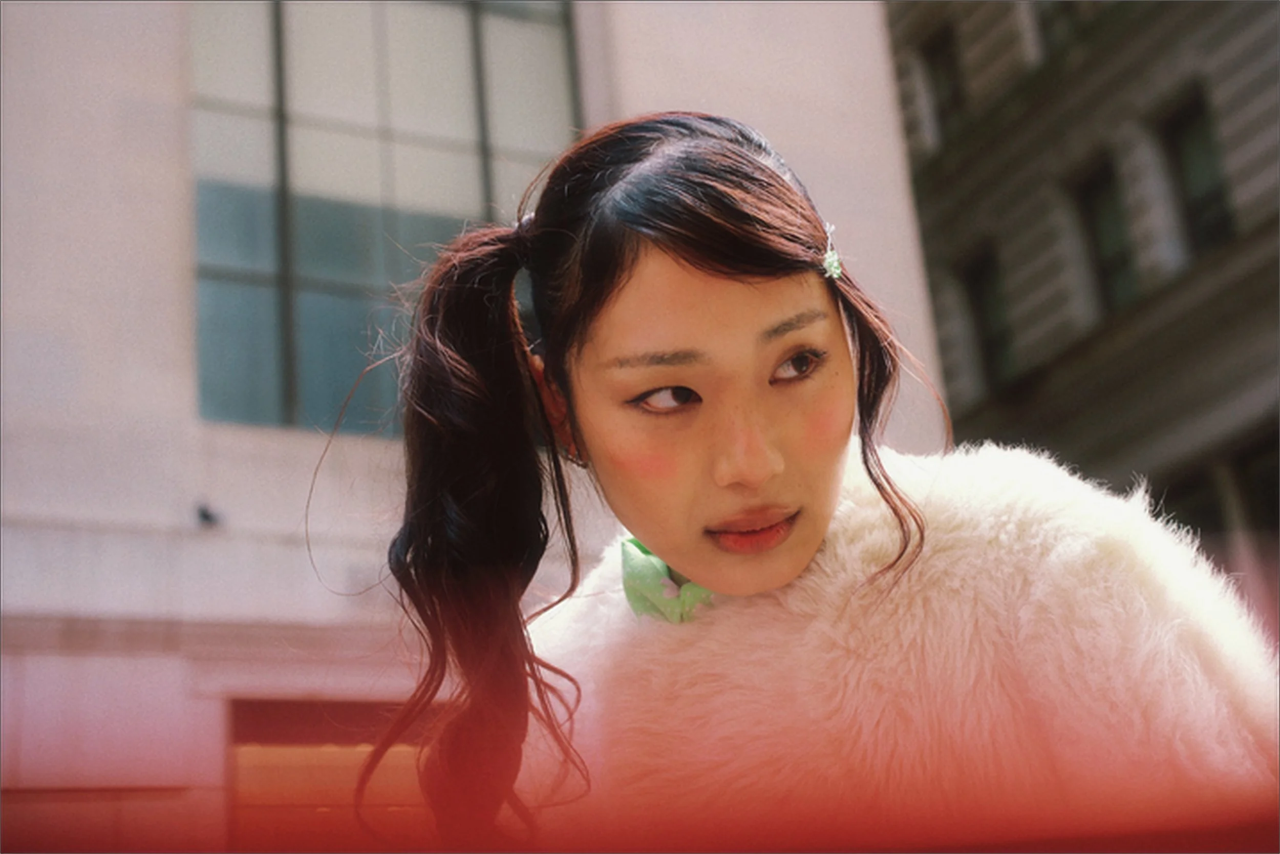 A woman with dark hair tied in pigtails, wearing a fluffy light-colored coat, with a green bow near her collar, standing outdoors with buildings in the background.