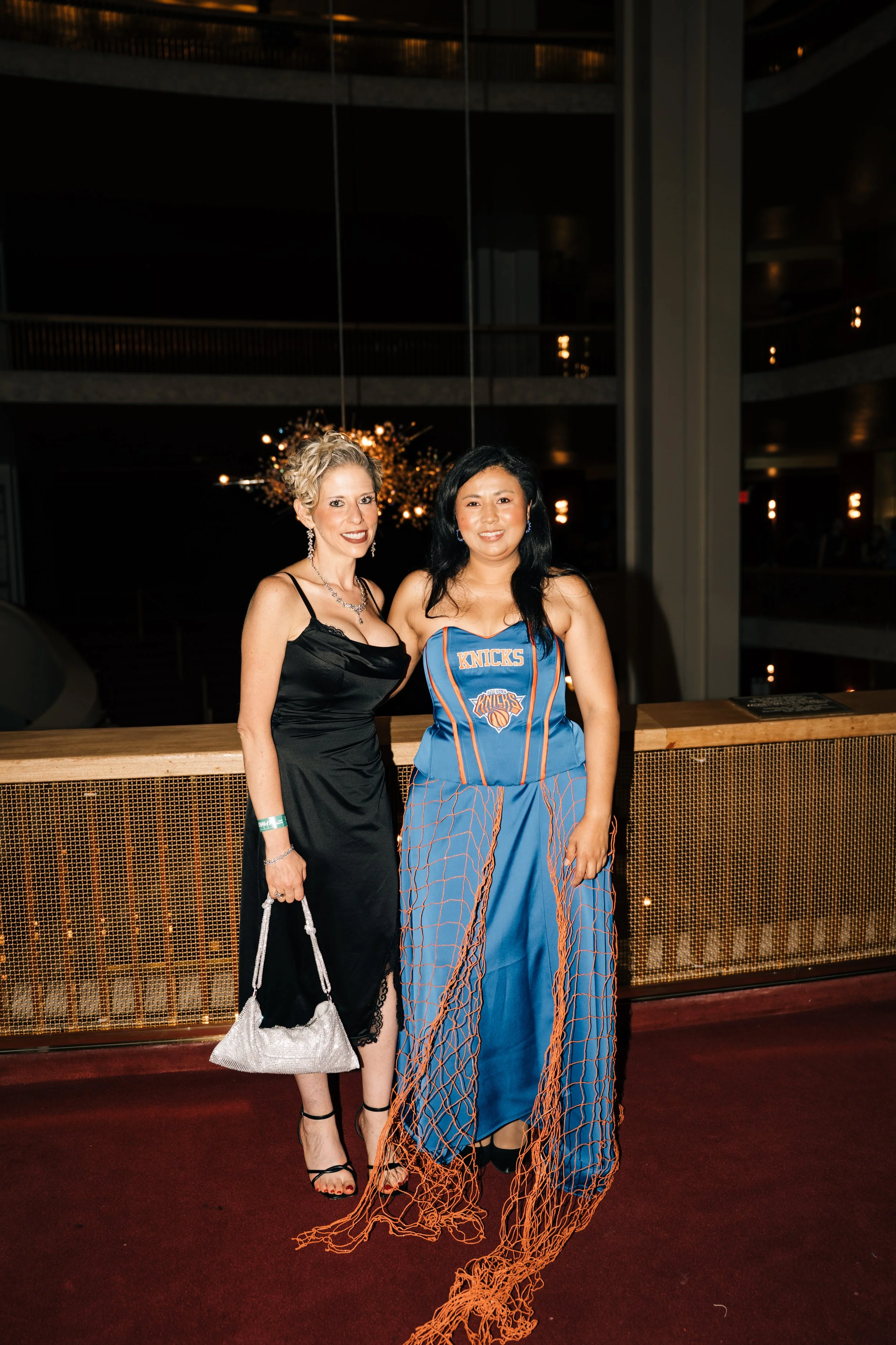 Two women posing together at a formal event. The woman on the left is wearing a black satin dress with lace detail and jewelry, holding a silver bag. The woman on the right is wearing a New York Knicks basketball jersey and matching mesh pants. They 